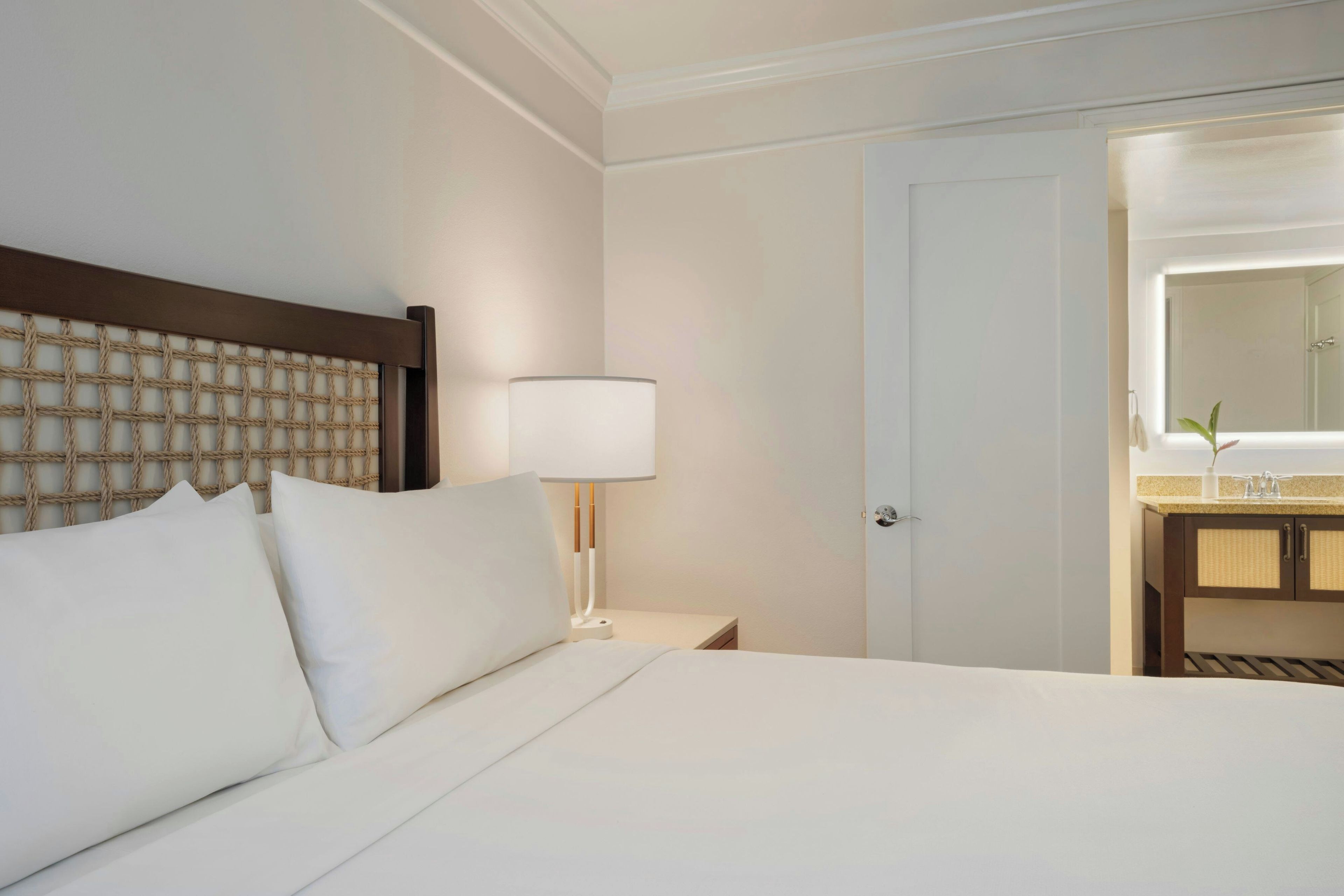 Una camera d'albergo con un letto, un comodino, una lampada, uno specchio e un bagno visibile attraverso una porta aperta.