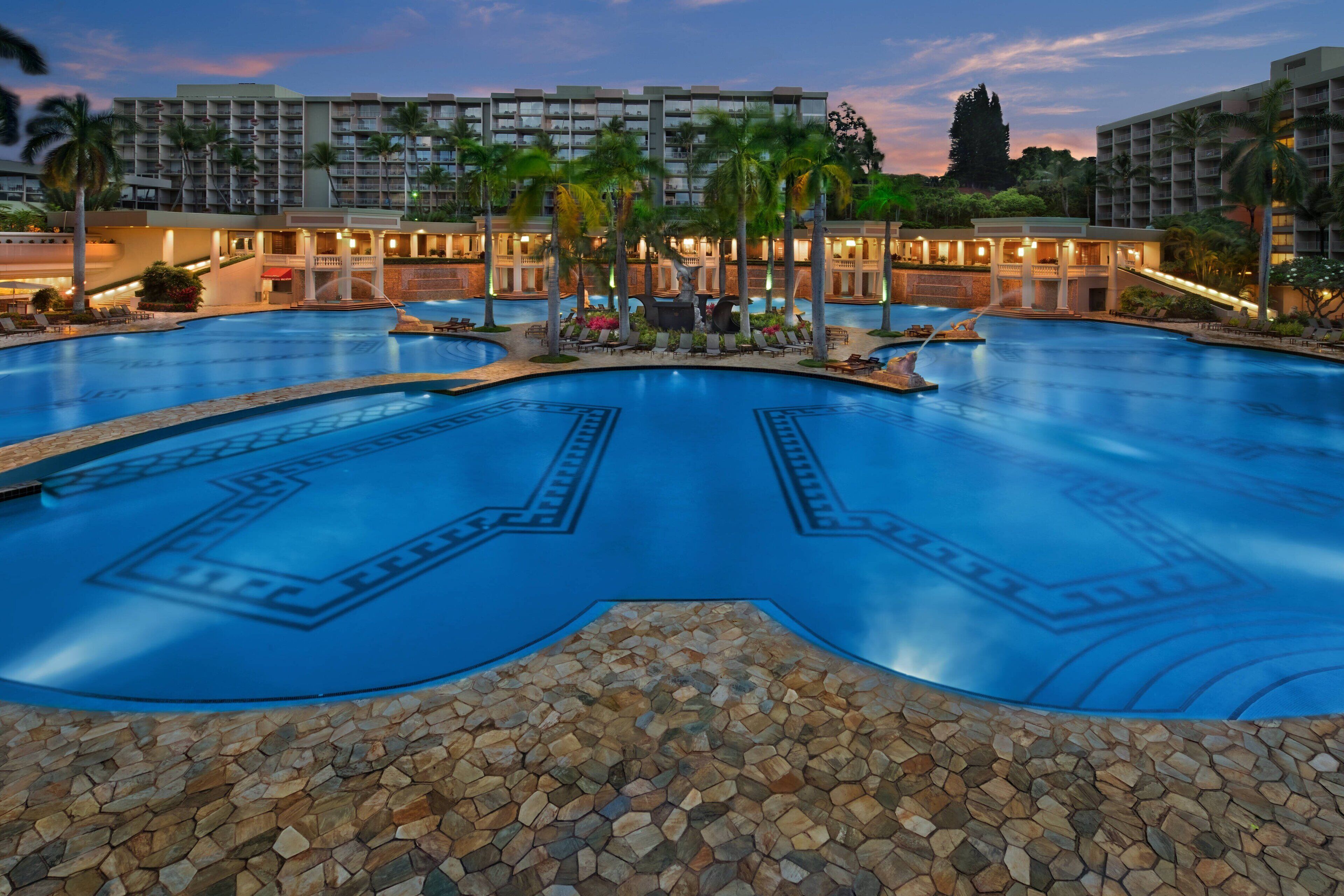A large swimming pool with a geometric design, surrounded by palm trees and a hotel building in the background.
