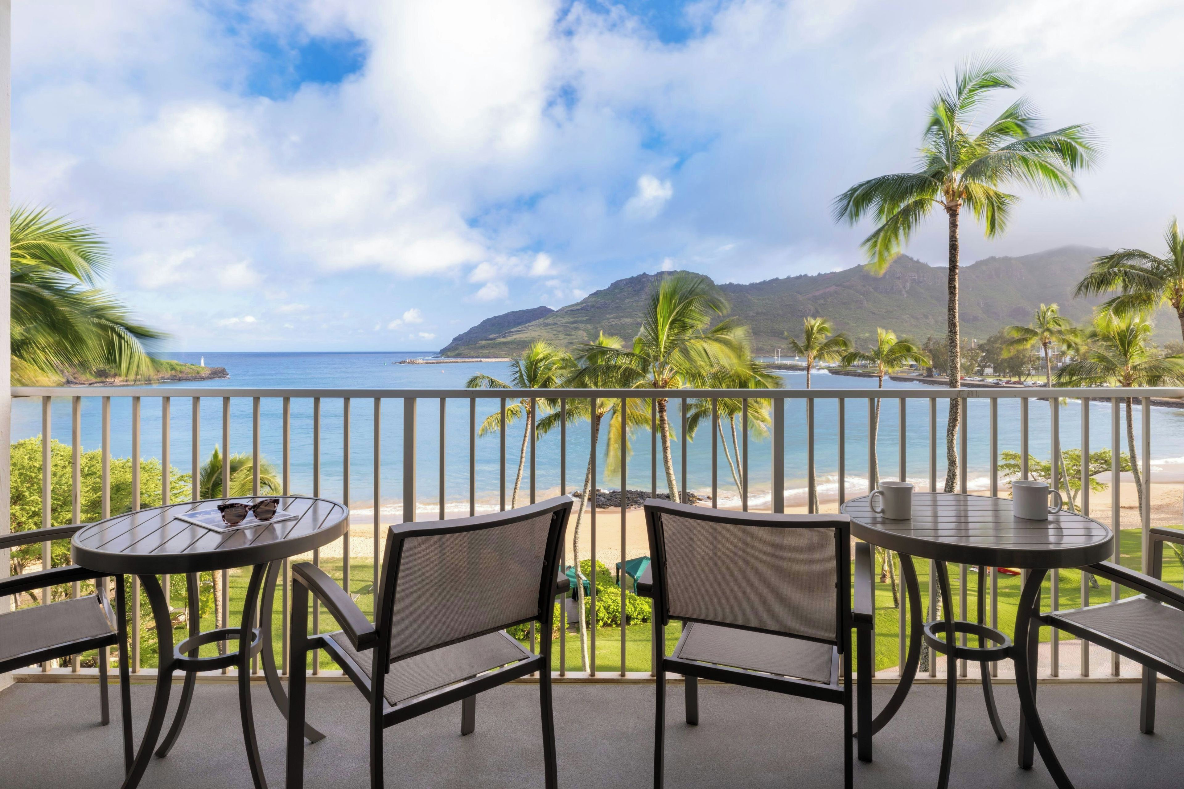 A balcony with two tables and chairs, overlooking a beach and a mountain.