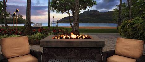 A fire pit with two wicker chairs, a stone table, and a view of the beach and ocean.