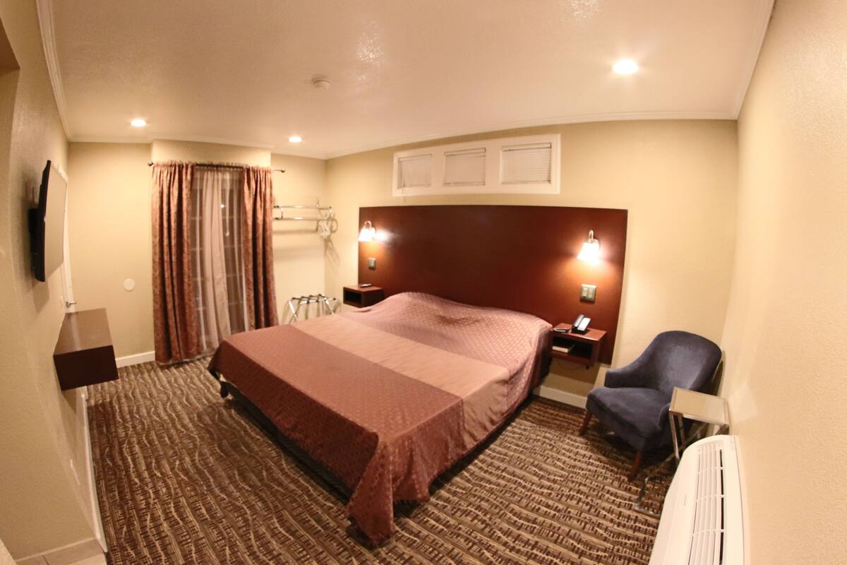 A hotel room with a large bed, a chair, a desk, a flat-screen TV, a window with curtains, and a wall-mounted light fixture.