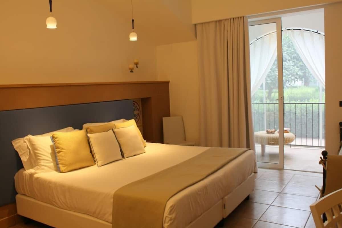 A bedroom with a large bed, a chair, a table, and a balcony with curtains.