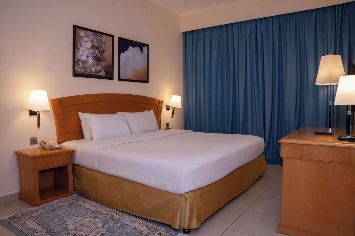 A hotel room with a large bed, two bedside lamps, a wooden nightstand, a desk, and a blue curtain.