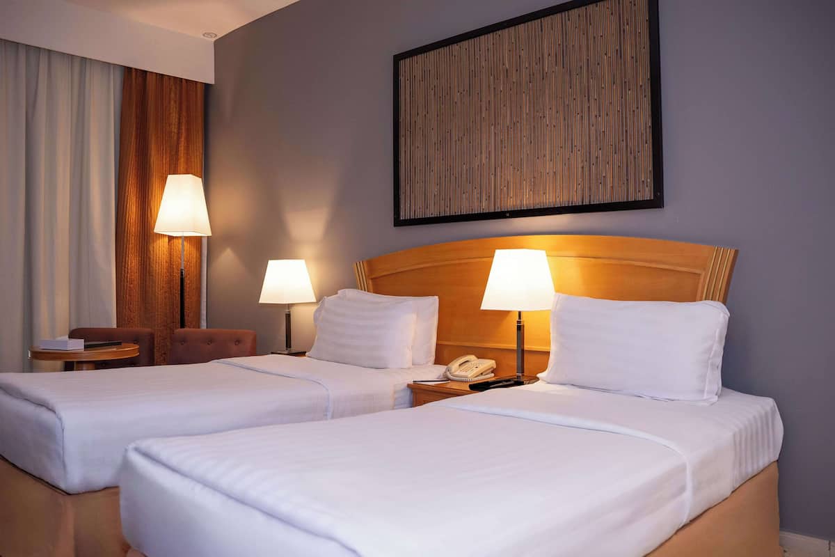 A hotel room with two beds, a wooden headboard, a bedside table with a lamp, a telephone, and a framed artwork on the wall.
