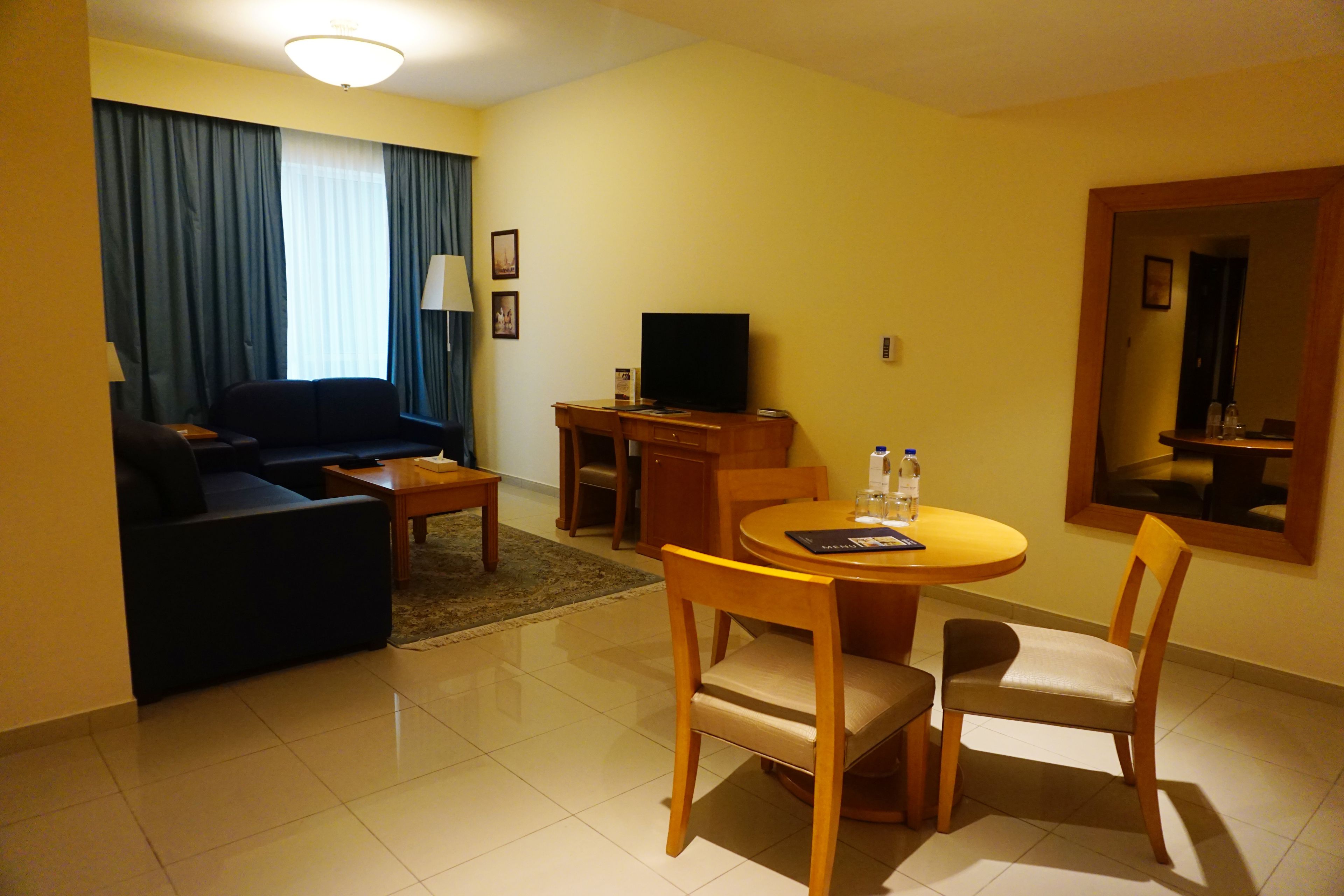A hotel room with a living area including a sofa, a TV, a dining table with chairs, and a small desk.