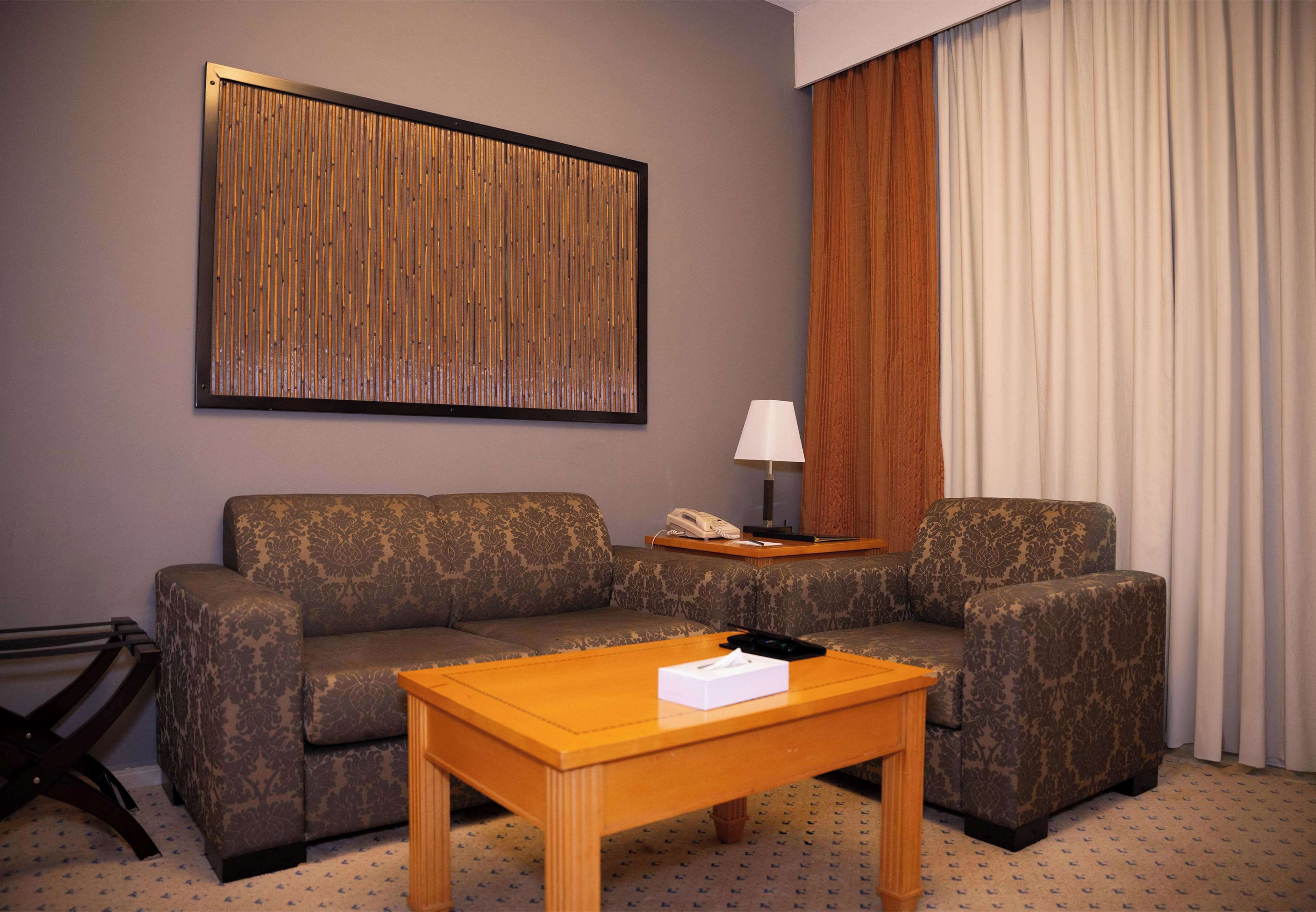 A hotel room with a sofa, a wooden coffee table, a lamp, and a framed artwork on the wall.