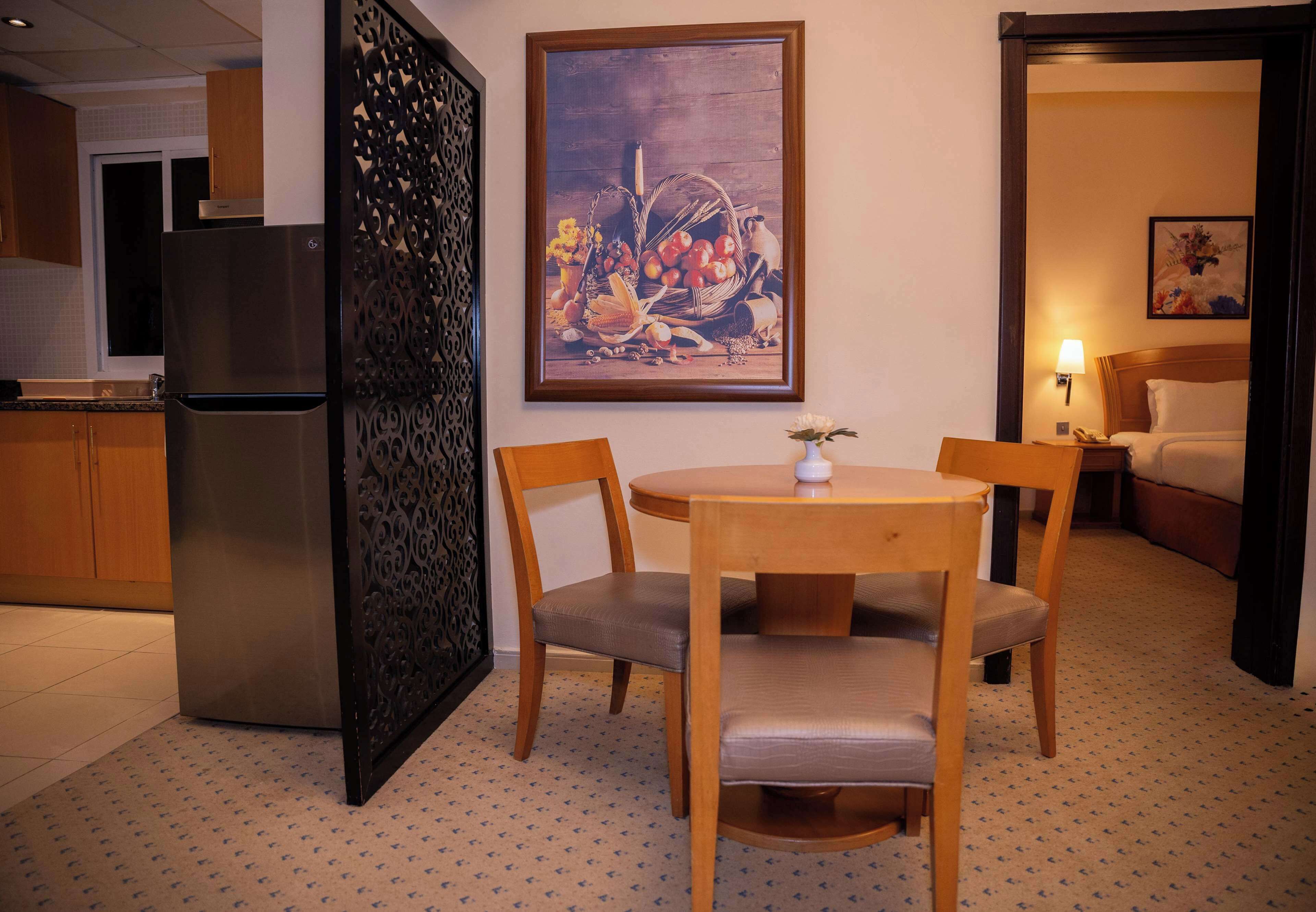 Ein Hotelzimmer mit Essbereich, ausgestattet mit einem Holztisch, Stühlen, einem Kühlschrank, einer Wanddekoration und einem Bett mit Nachttisch.