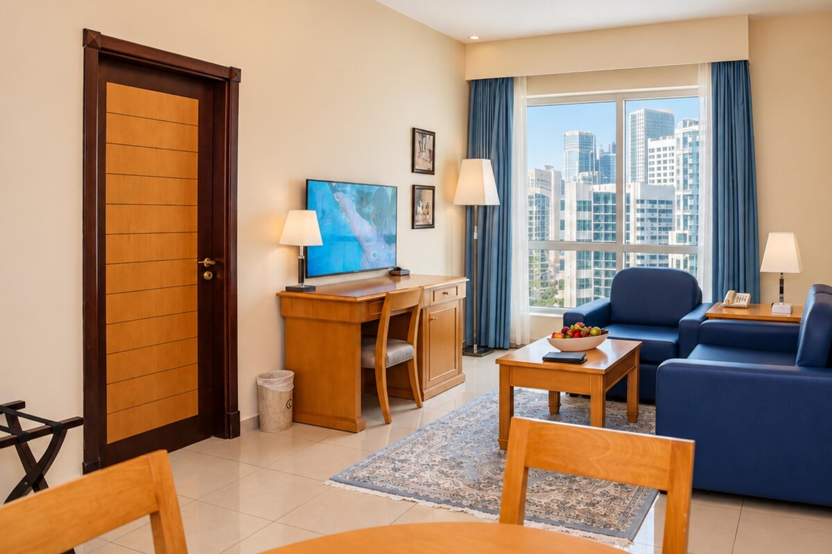 A hotel room with a desk, a blue sofa, a television, and a view of the cityscape.