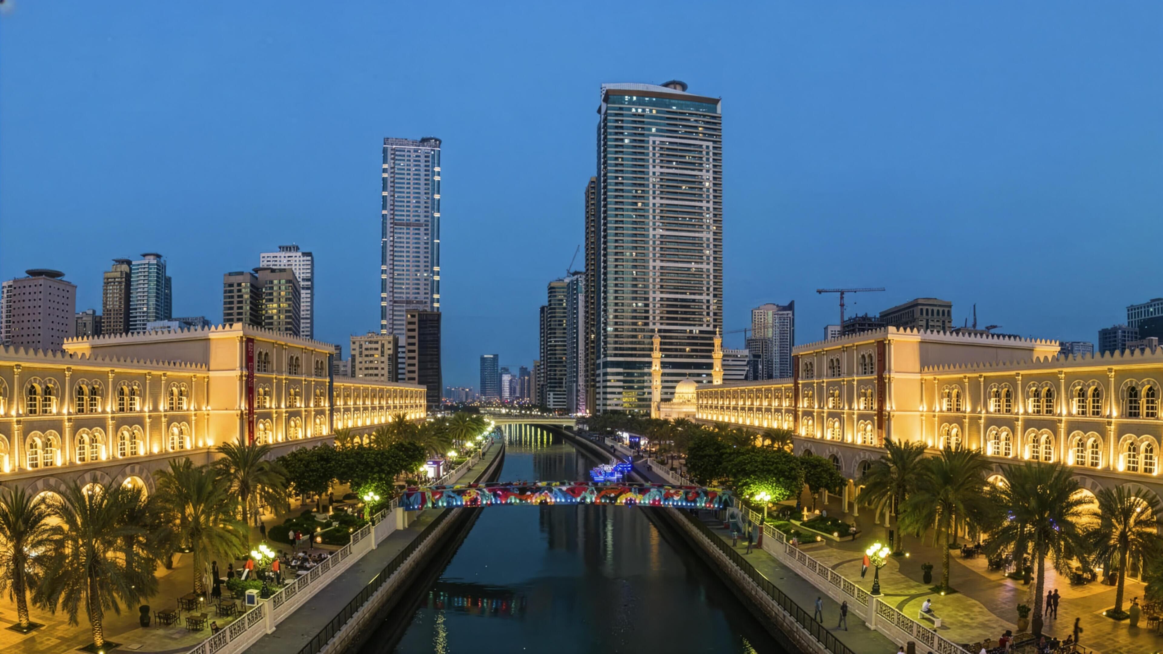 Un paisaje urbano con un río en primer plano, flanqueado por dos edificios históricos con ventanas arqueadas y un rascacielos moderno al fondo.