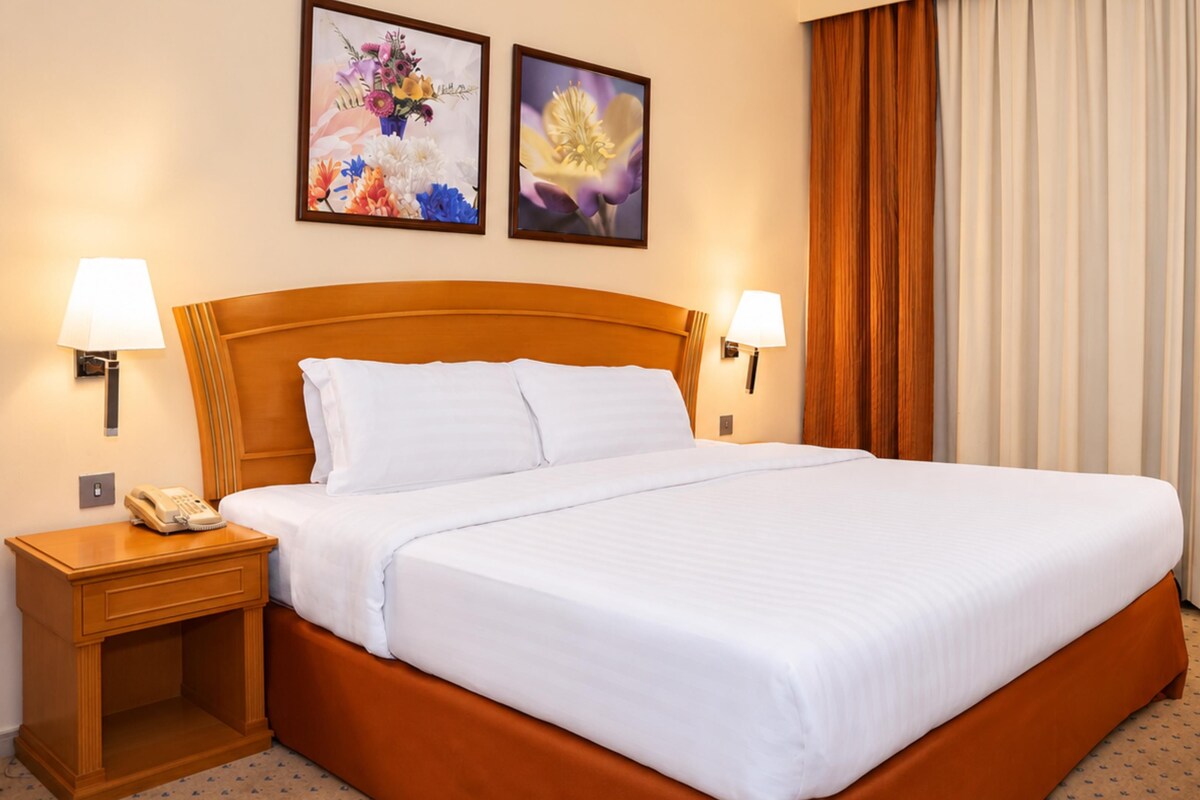 A hotel room with a large bed, two bedside lamps, a telephone, and two framed floral pictures on the wall.