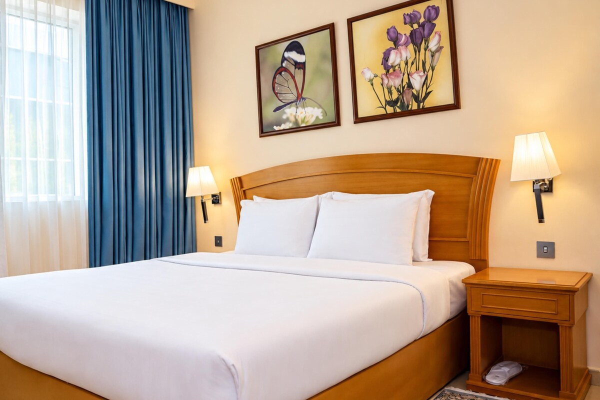 A hotel room with a large bed, two bedside lamps, a nightstand, a window with curtains, and two wall-mounted paintings.