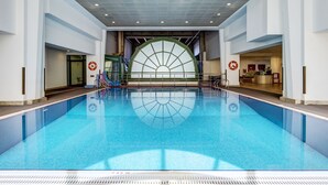 Una amplia piscina interior con agua cristalina azul, rodeada de paredes blancas y un diseño moderno de techo.