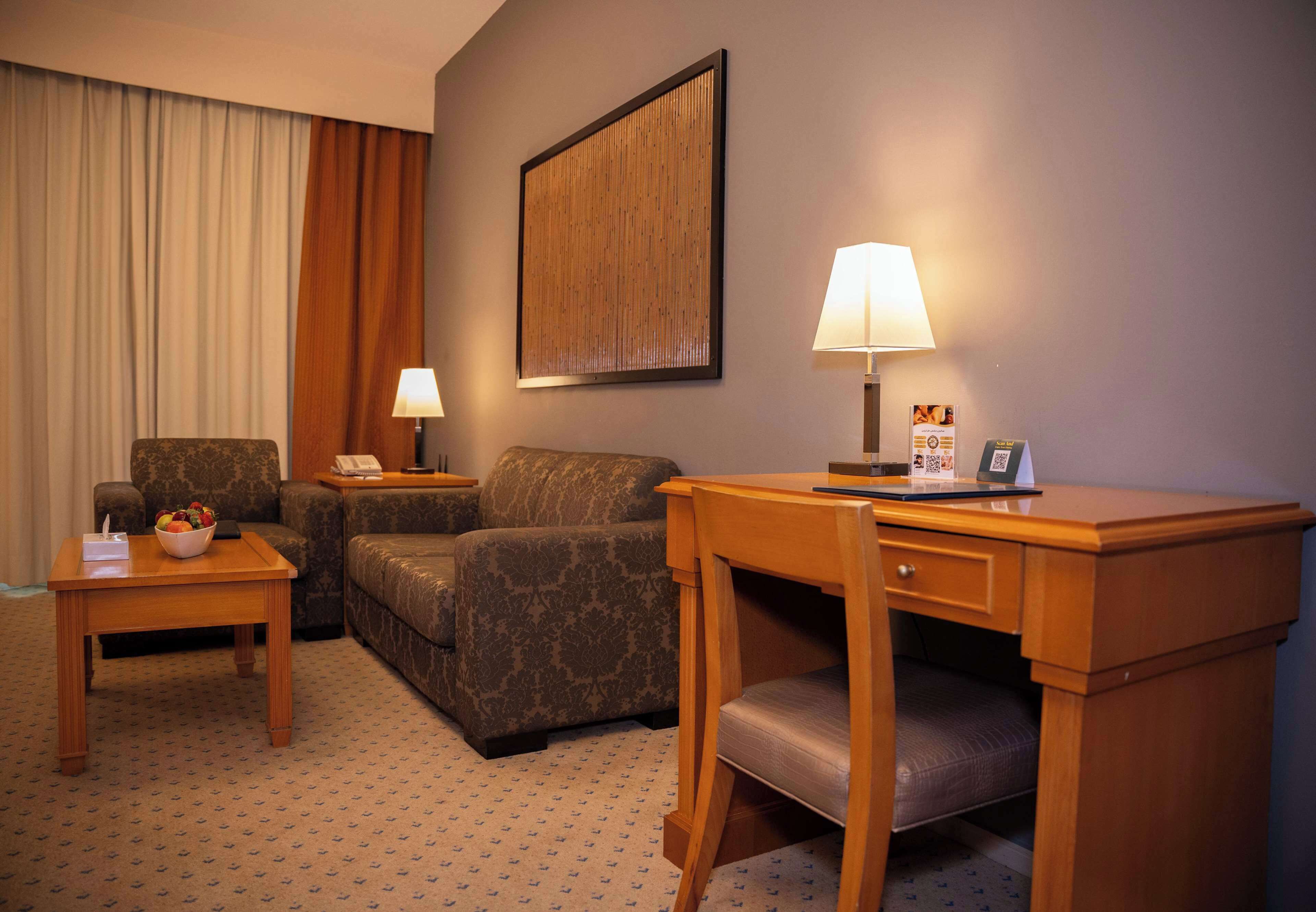 A hotel room with a sofa, two side tables, a desk with a chair, a lamp, and a framed picture on the wall.