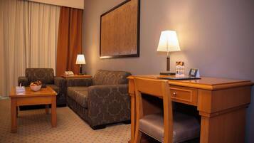 A hotel room with a sofa, two side tables, a desk with a chair, a lamp, and a framed picture on the wall.