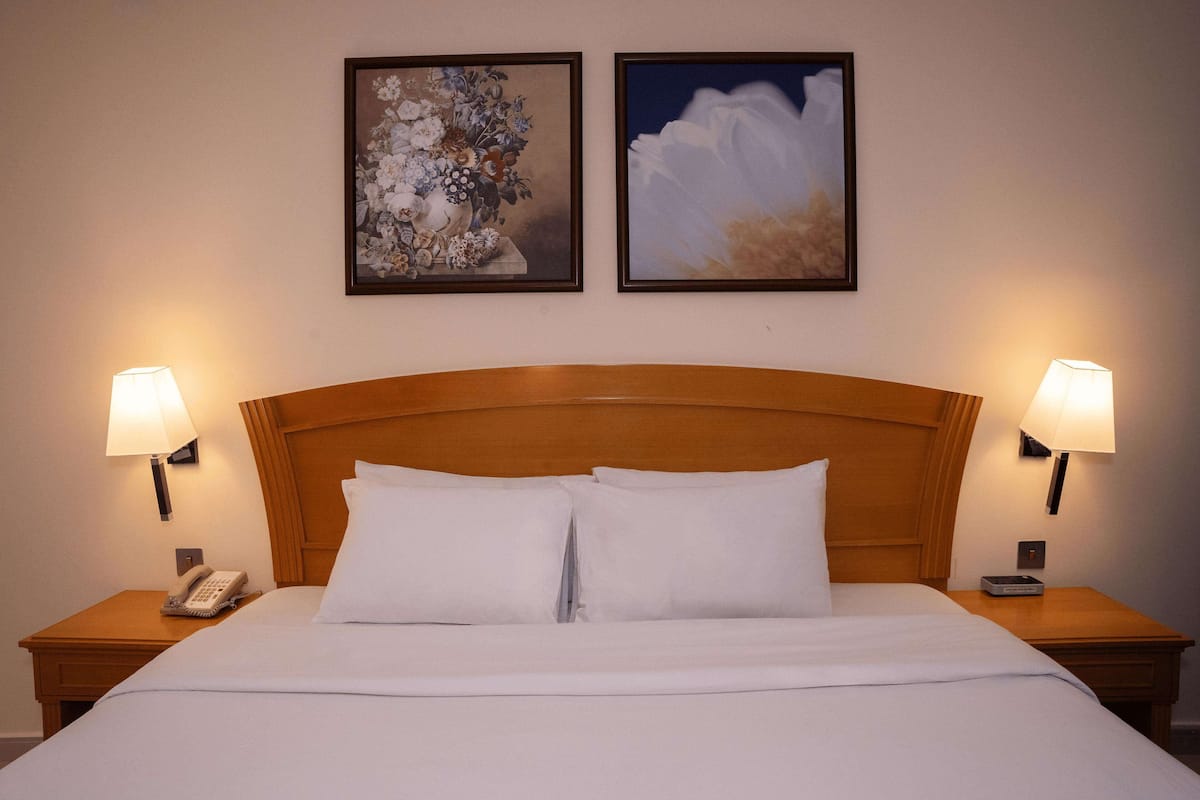 A hotel room with a wooden headboard, two bedside lamps, a telephone, and two framed floral paintings on the wall.