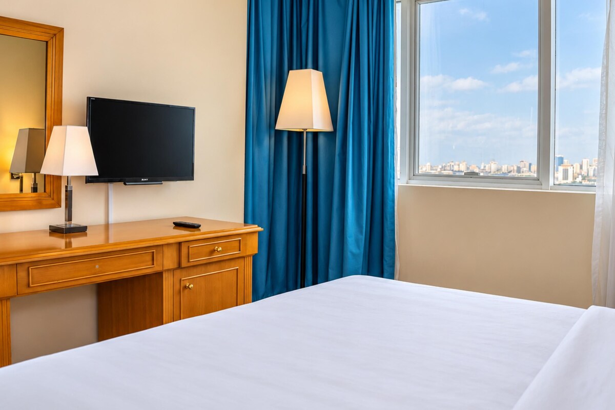 A hotel room with a bed, a desk with a lamp, a television, and a window with curtains.