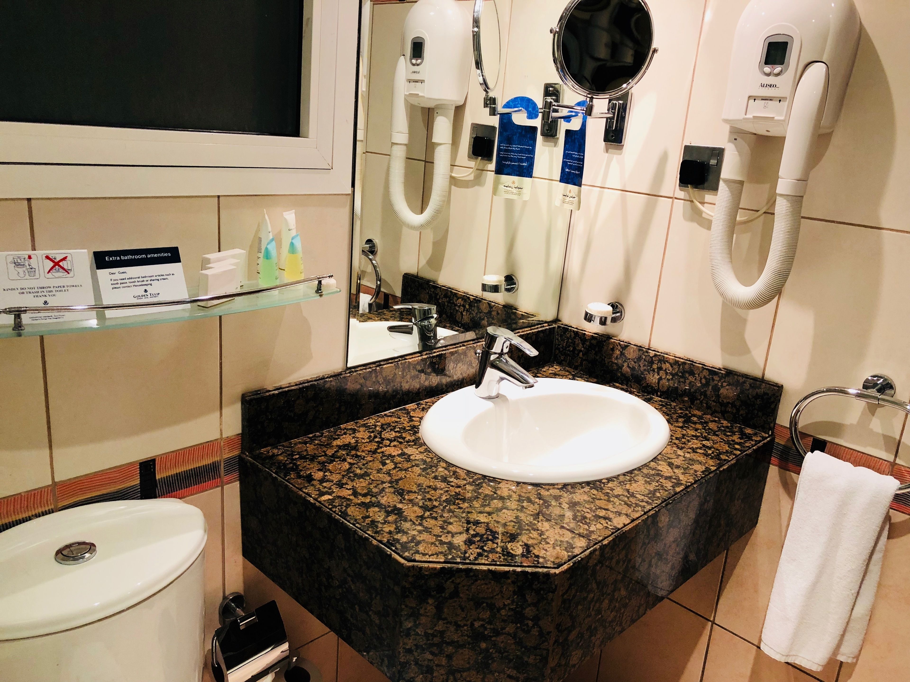 A bathroom with a granite countertop sink, a mirror, a towel rack, and a shelf with toiletries.