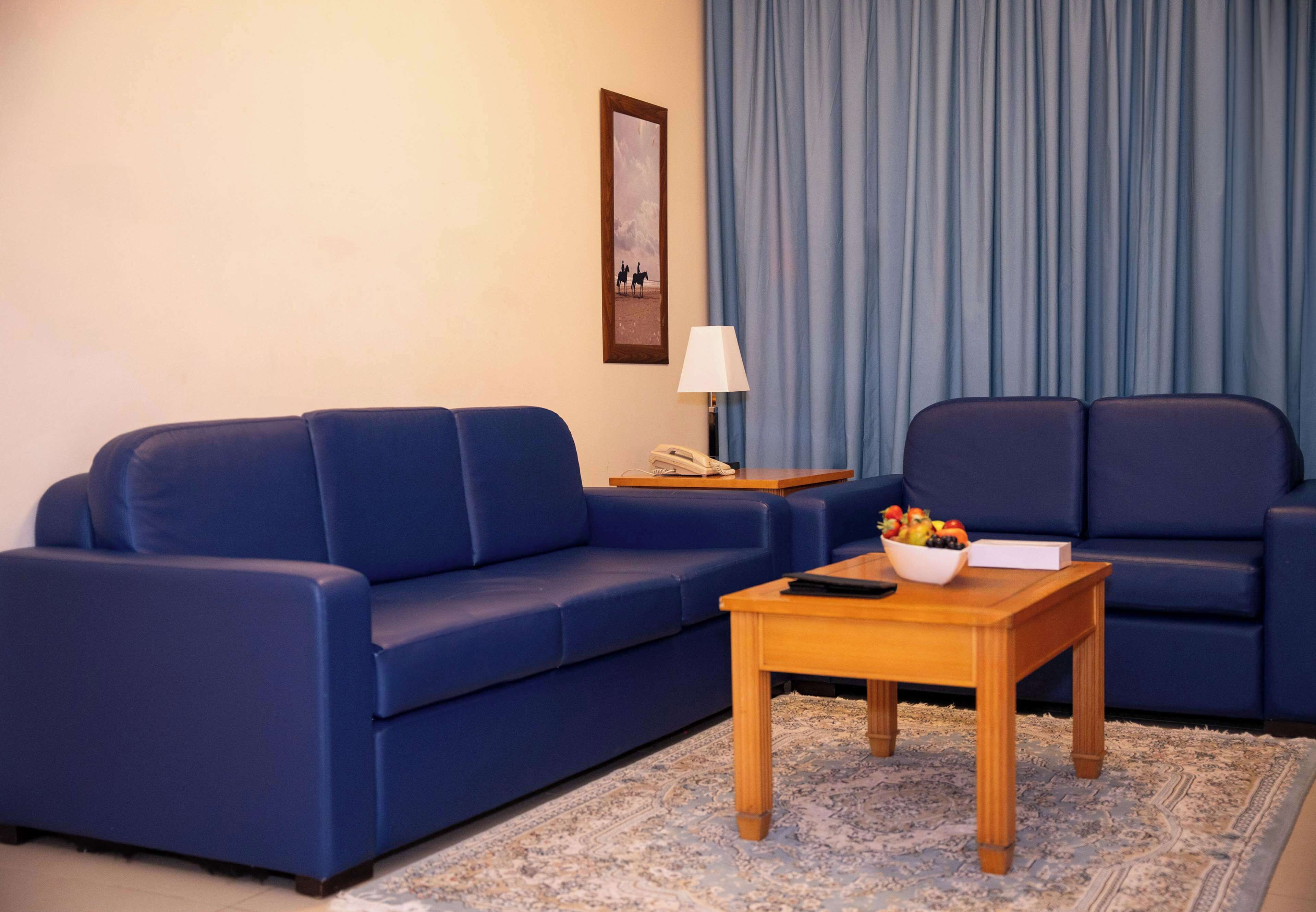 A living room with a blue sofa, a wooden coffee table with a bowl of fruit, a lamp, and a framed picture on the wall.
