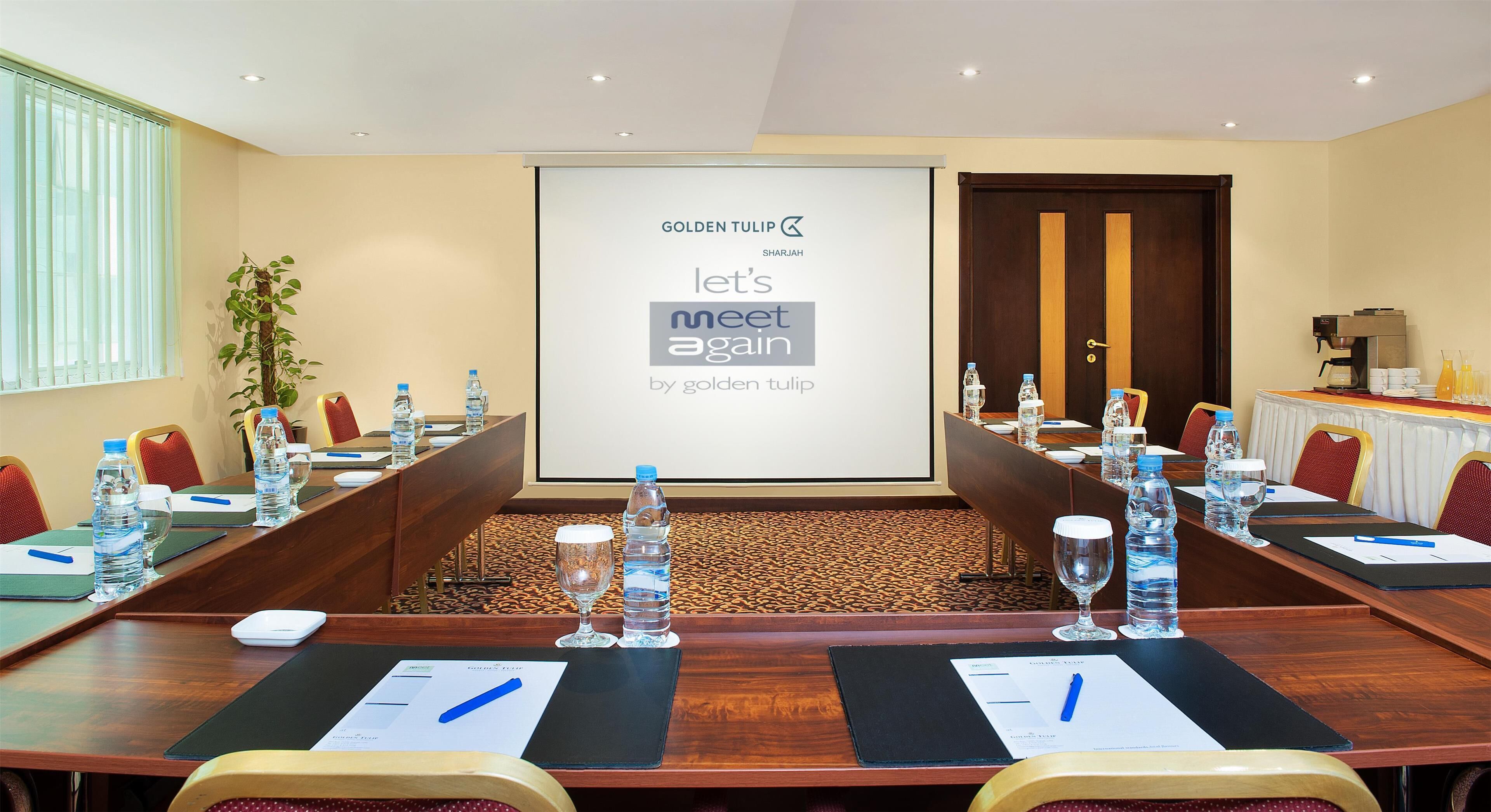A conference room with a long table, water bottles, and a projector screen displaying "GOLDEN TULIP SHARJAH let's meet again by golden tulip".