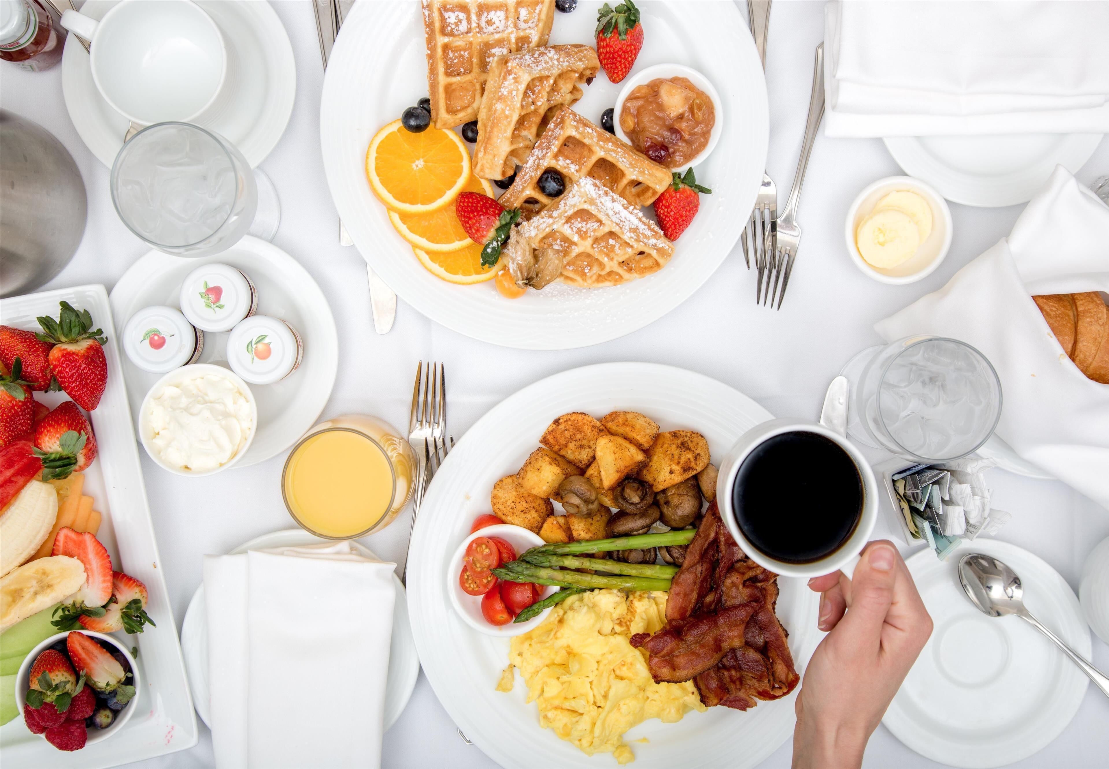 A breakfast table with waffles, strawberries, blueberries, orange slices, scrambled eggs, bacon, asparagus, and a cup of coffee.