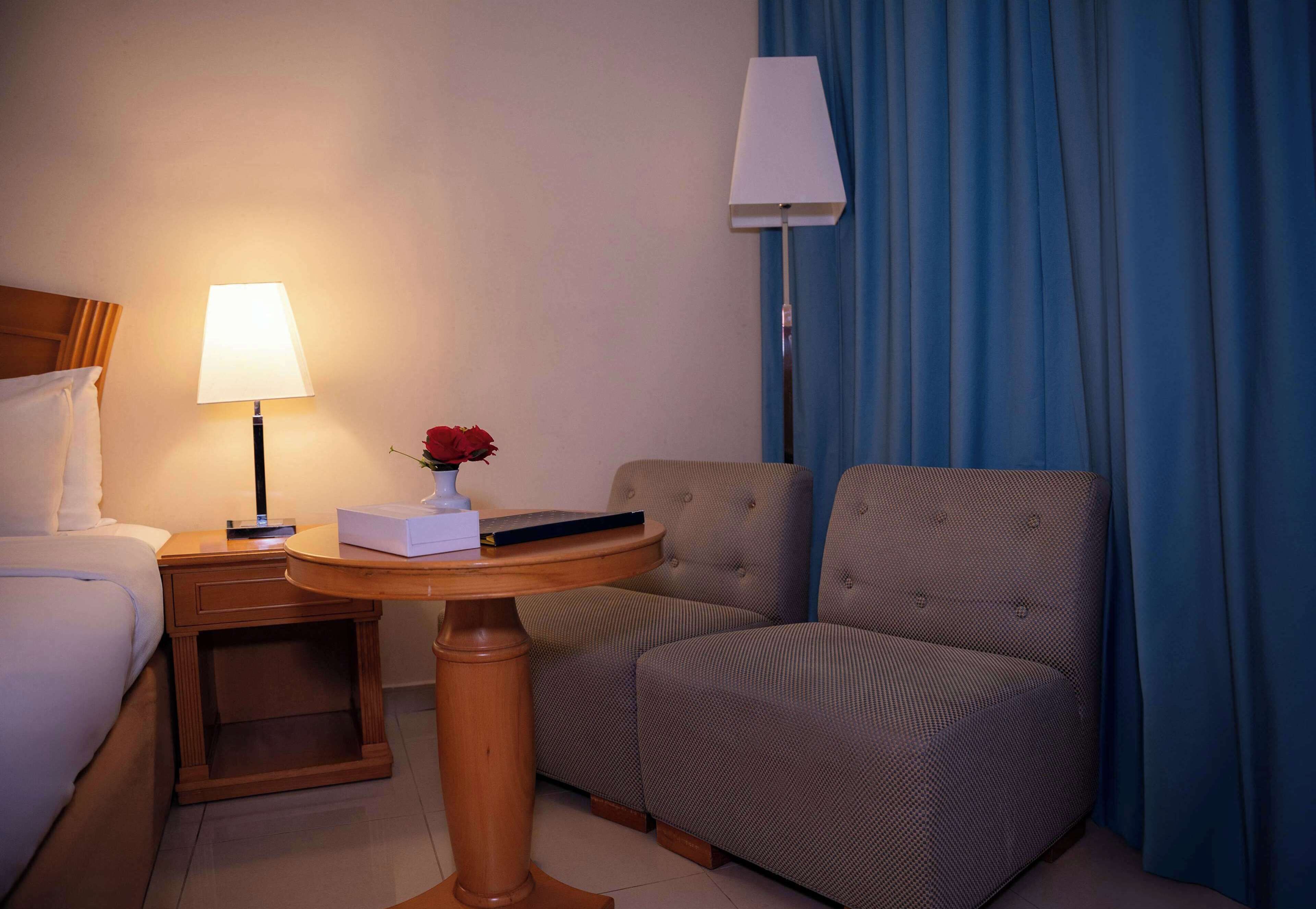 A hotel room with a bed, a bedside table with a lamp, a small round table with a vase of flowers, and a chair.