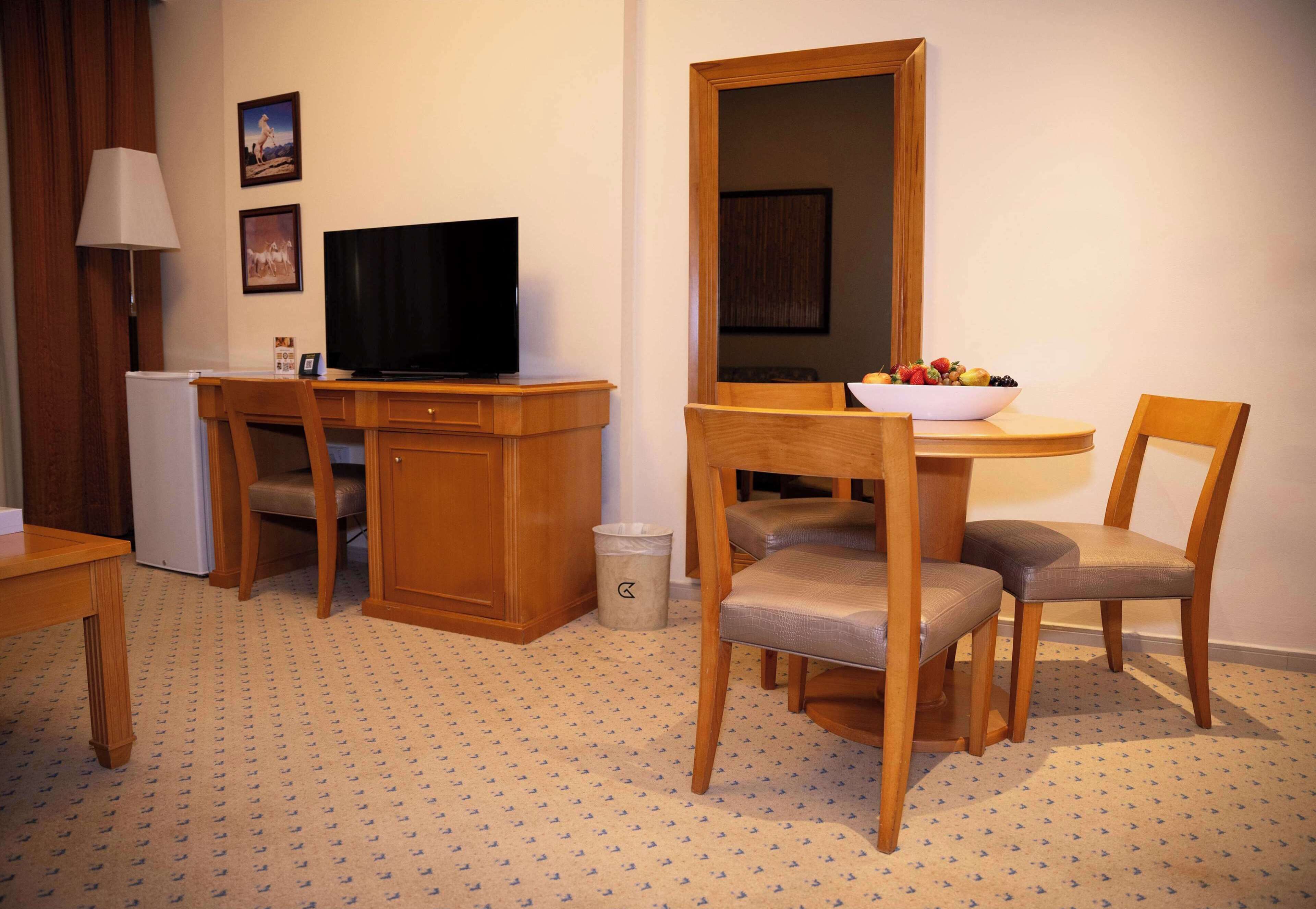 Ein Hotelzimmer mit einem Holztisch, einem Fernseher, einem kleinen Kühlschrank, einem Mülleimer und einem Essbereich mit einer Fruchtschale und zwei Stühlen.