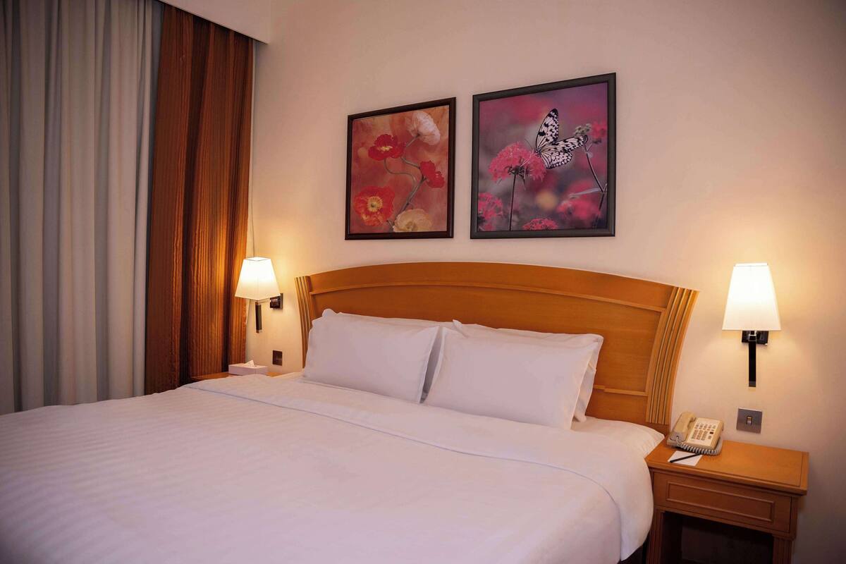 A hotel room with a large bed, two wall-mounted lamps, a nightstand with a phone, and two framed floral paintings on the wall.