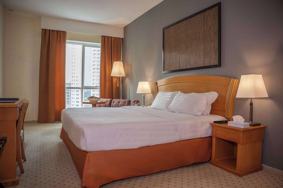 A hotel room with a large bed, two bedside lamps, a wooden nightstand, a desk with a chair, and a view of buildings through the window.