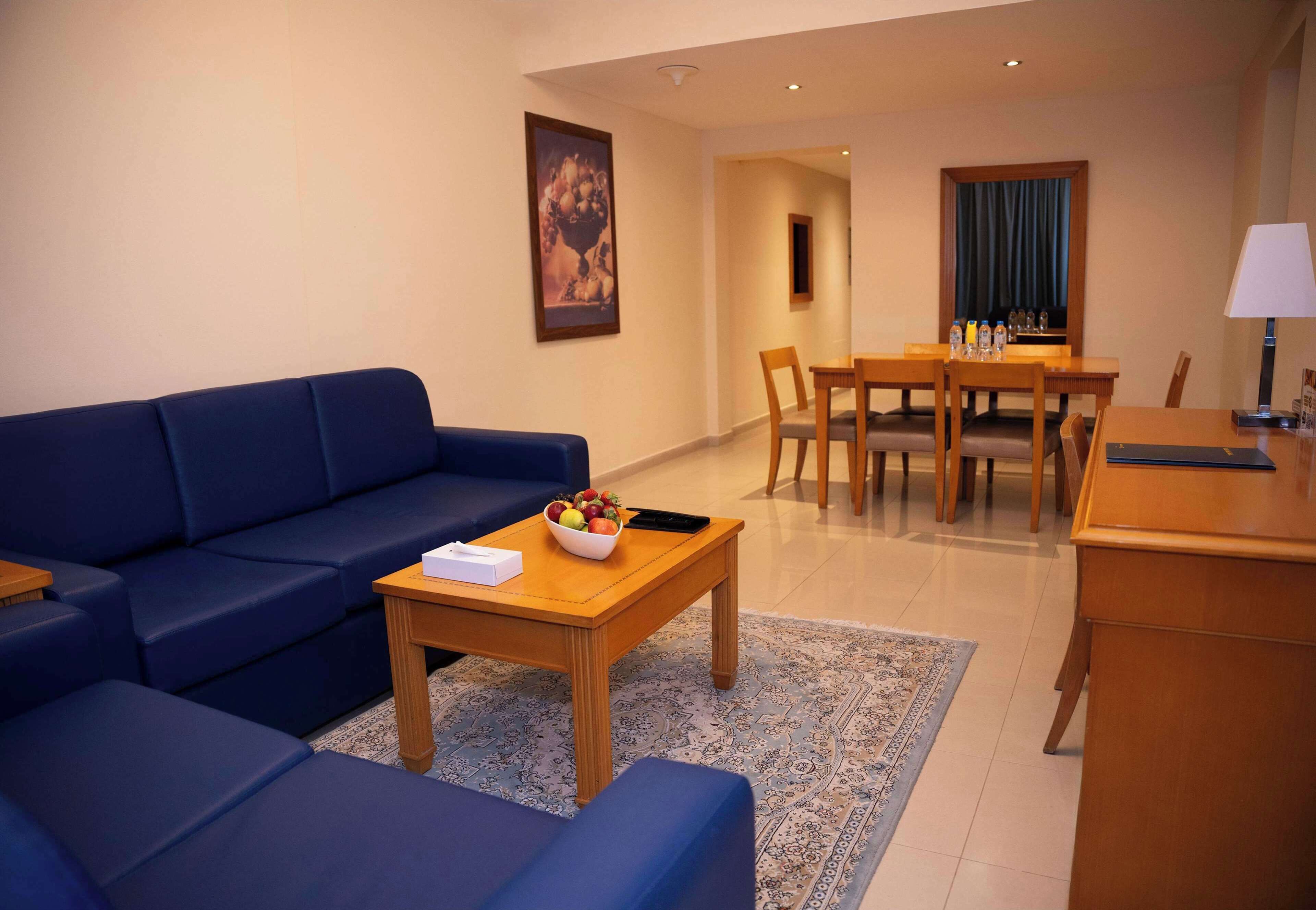 A modern hotel room with a blue sofa, a wooden coffee table with a fruit bowl, a dining area with a table and chairs, and a desk with a lamp.