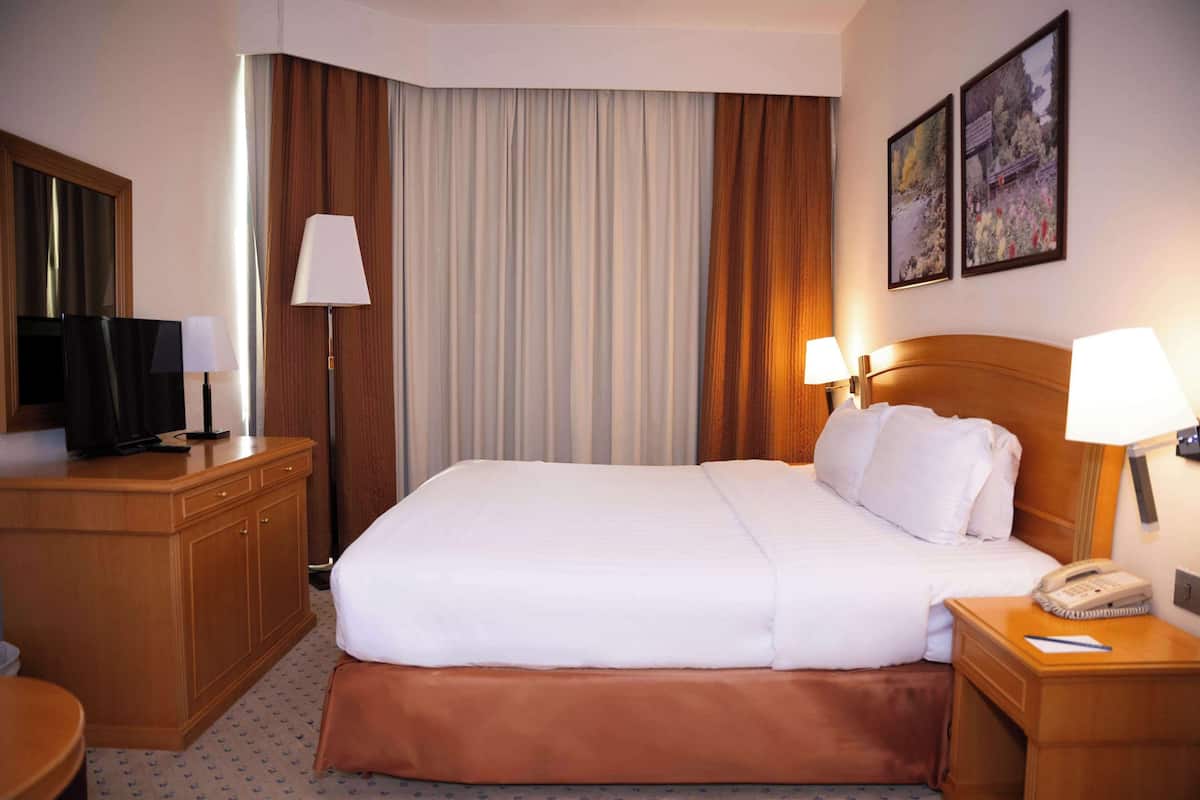 A hotel room with a large bed, wooden headboard, two bedside lamps, a desk with a telephone, and a television on a wooden cabinet.
