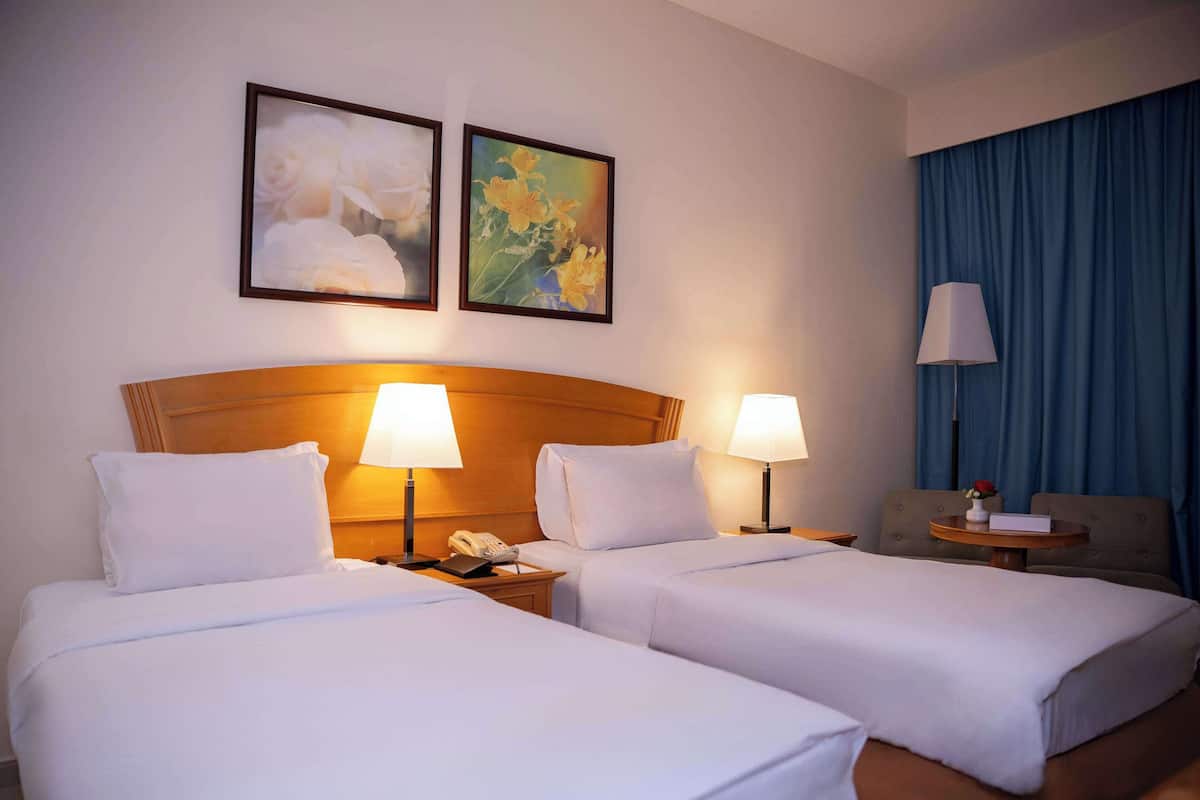 A hotel room with two beds, a wooden headboard, bedside lamps, a telephone, a small table with a vase, and two framed floral paintings on the wall.