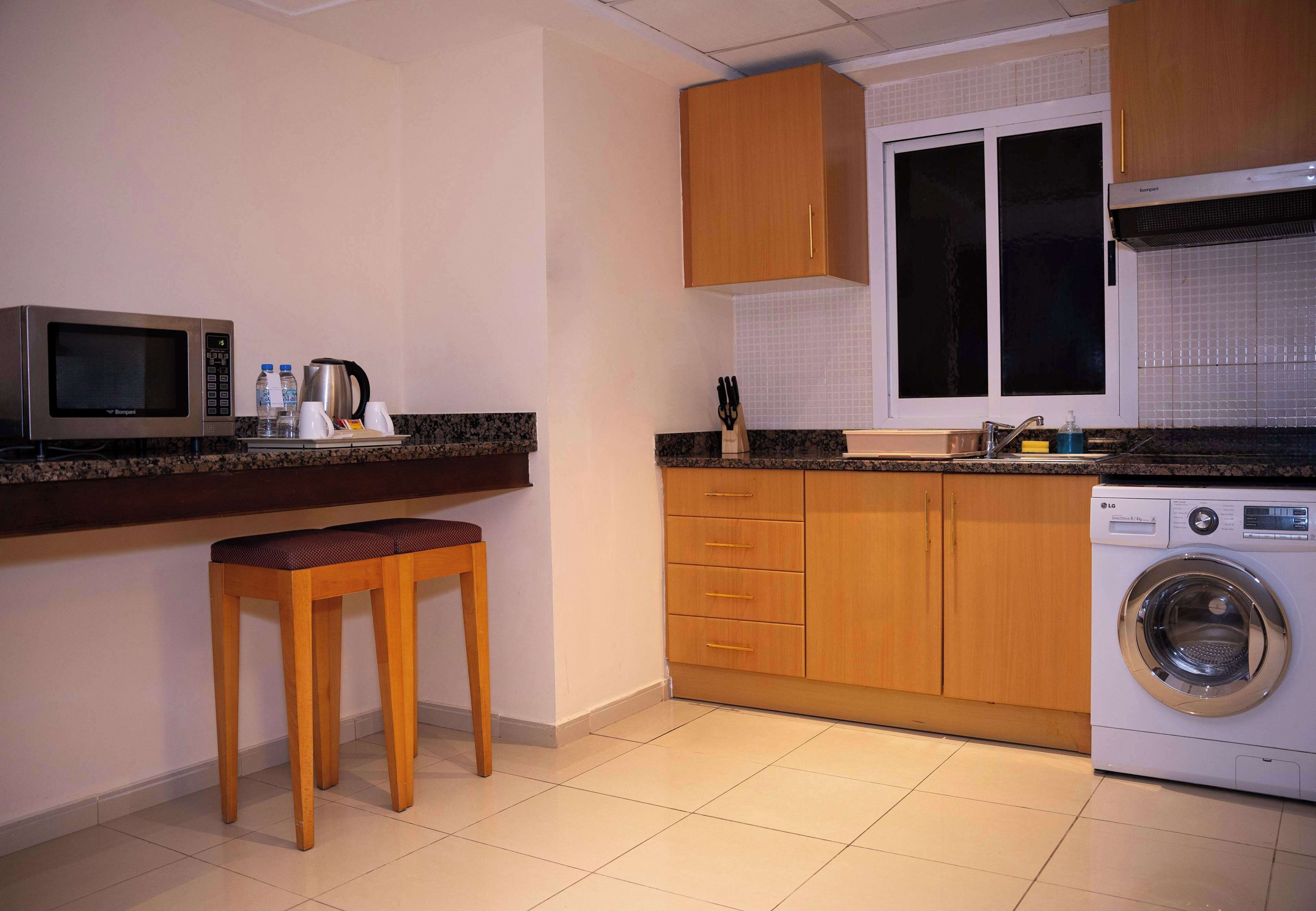 A compact kitchen with a microwave, coffee maker, and washing machine. There are wooden cabinets and a window above the sink.