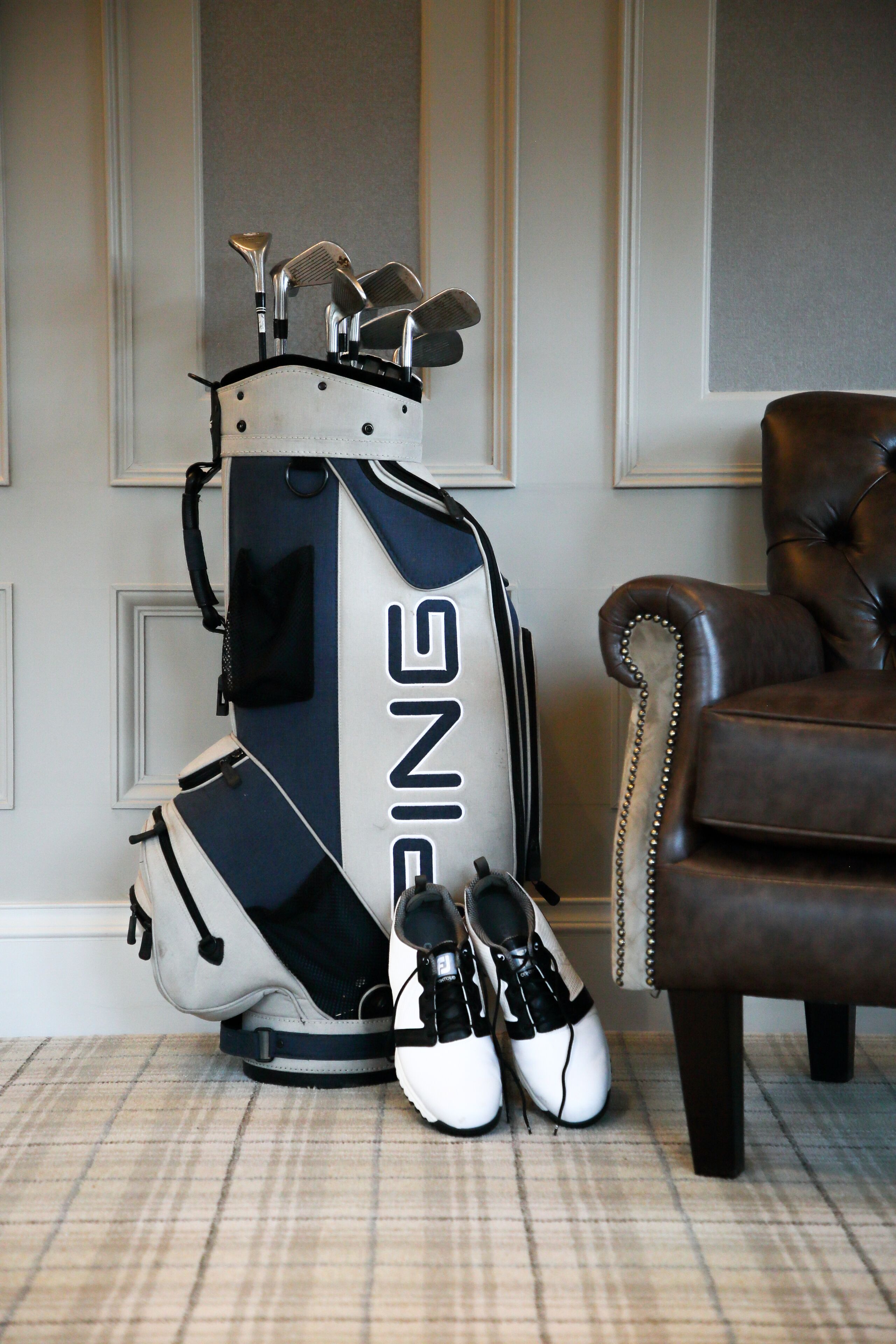 A golf bag with shoes beside it, a brown leather chair, and a checkered floor.