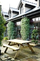Una mesa de picnic de madera frente a un edificio con grandes ventanales y un área pavimentada con piedra.