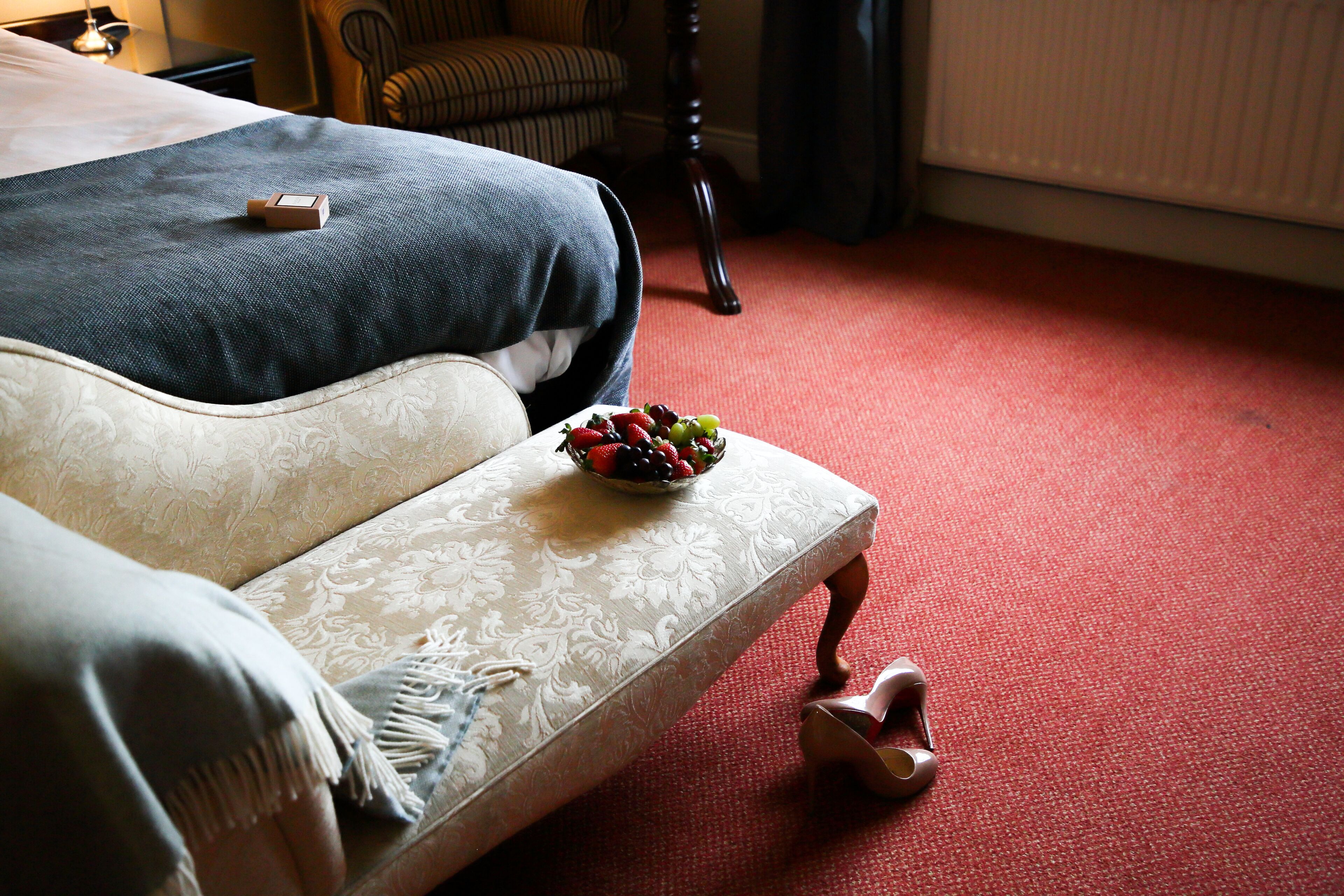 A neatly made bed with a blue blanket, a bedside table with a bowl of fruit, and a pair of shoes on the floor.