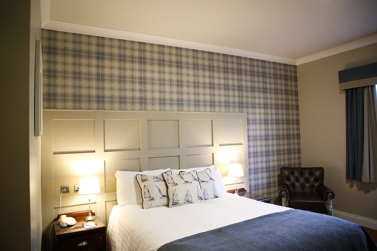 Una habitación de hotel con una cama grande, un cabecero estampado en pata de gallo, una mesita de noche con lámpara y una silla.