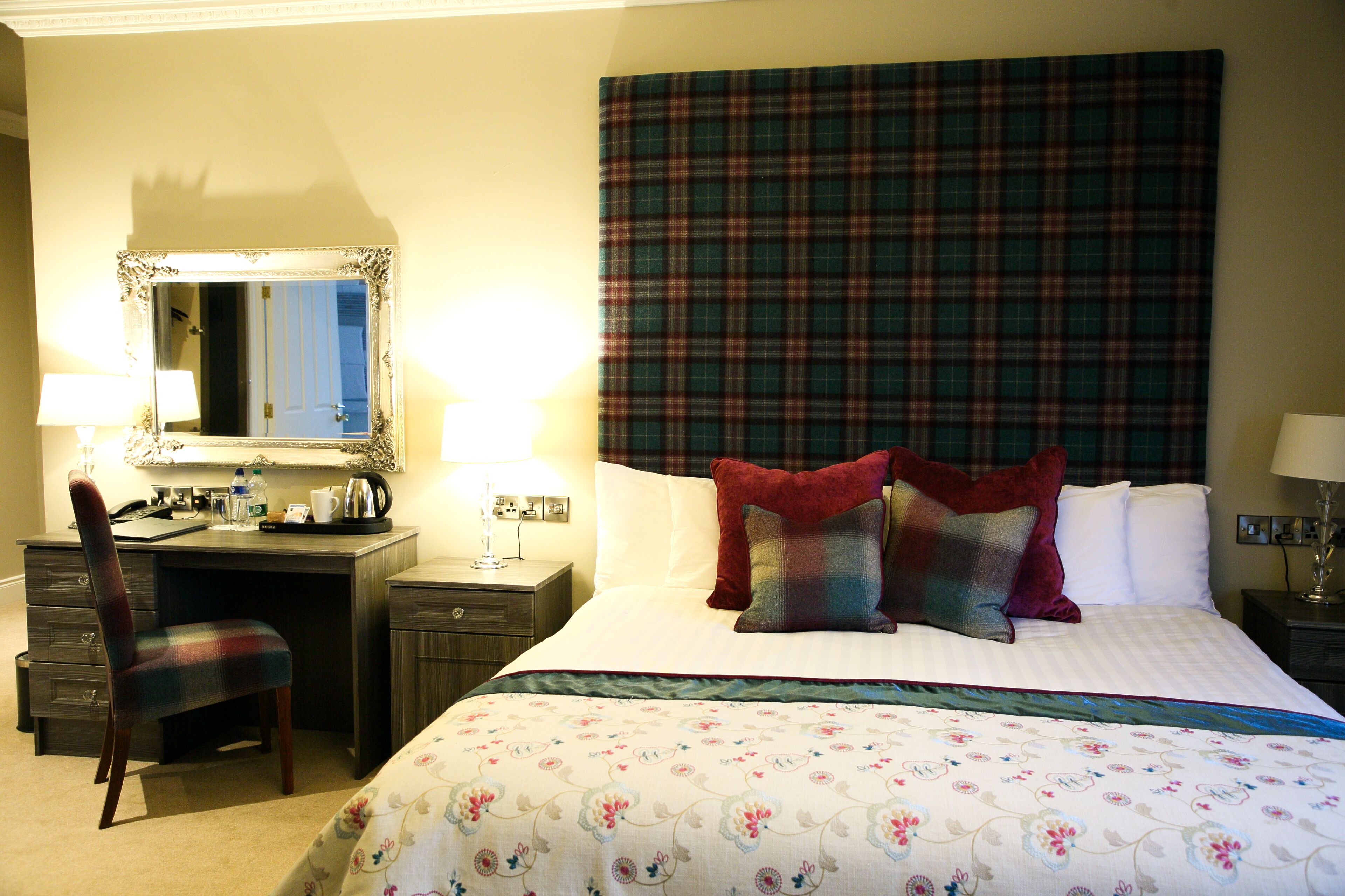 A hotel room with a bed, a desk, a chair, a mirror, and a plaid headboard.