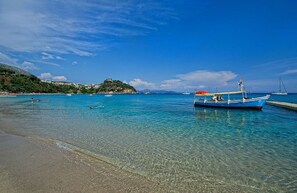 Ein unberührter Strand mit einem kleinen Boot, das an der Küste liegt, ruhiges Wasser und eine entfernte Hügelkette mit Gebäuden.