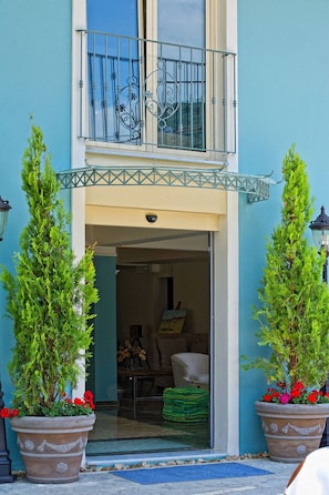 A building entrance with a glass door, a balcony with metal railings, and two potted plants with red flowers. - Parga Beach Resort (Parga)