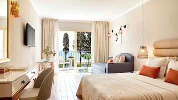 Habitación de hotel con cama, escritorio, silla, sofá, televisor y balcón con vista a árboles y al cielo.