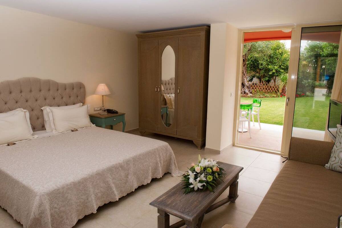 A bedroom with a large bed, a wooden wardrobe, a bedside table with a lamp, and a view of a garden through a sliding glass door.
