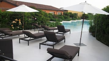 Zona junto a la piscina con tumbonas y sombrillas, rodeada de vegetación y un edificio con tejado de tejas.