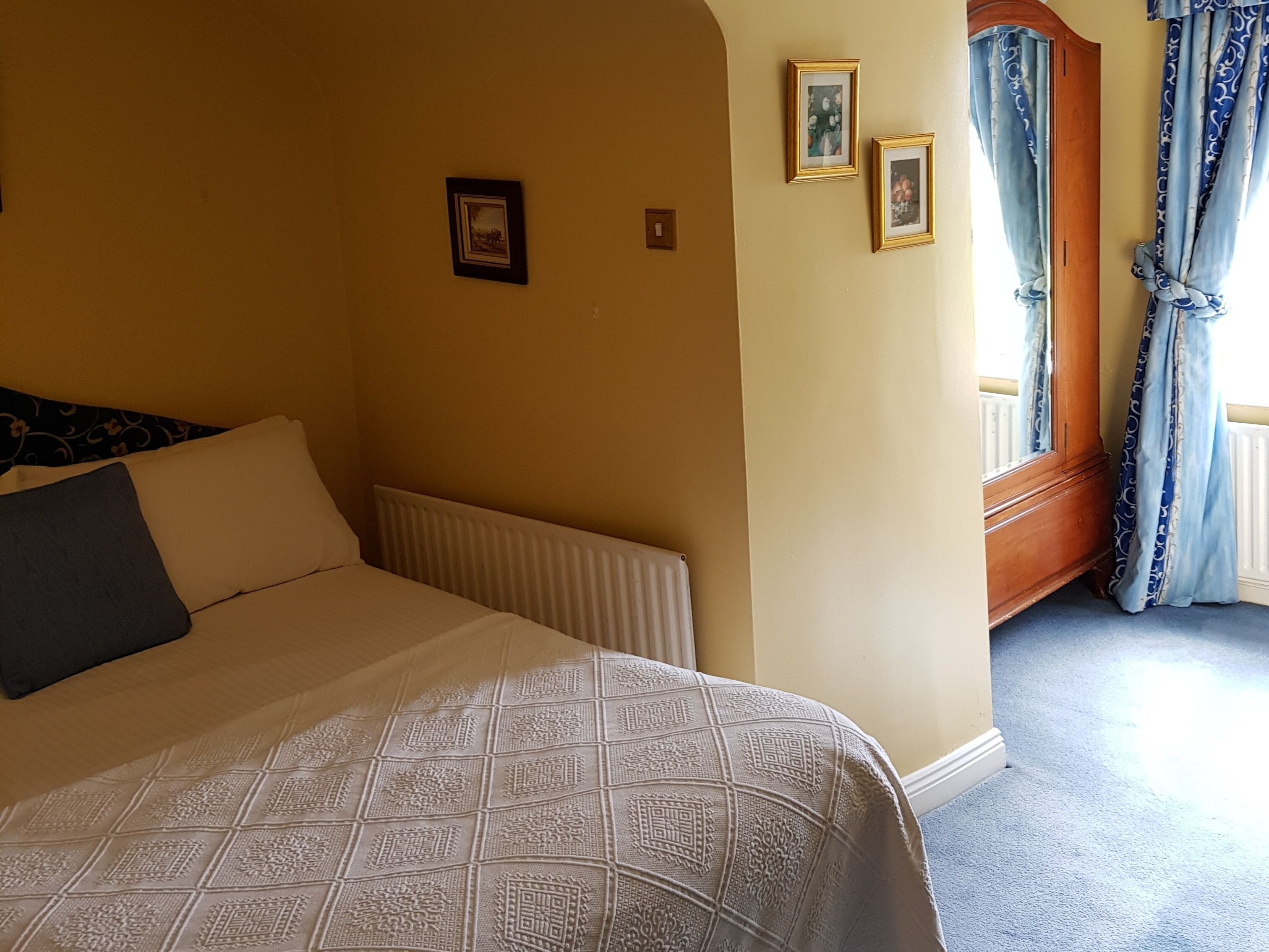 A hotel room with a large bed, a radiator, a wooden wardrobe, and a window with curtains.