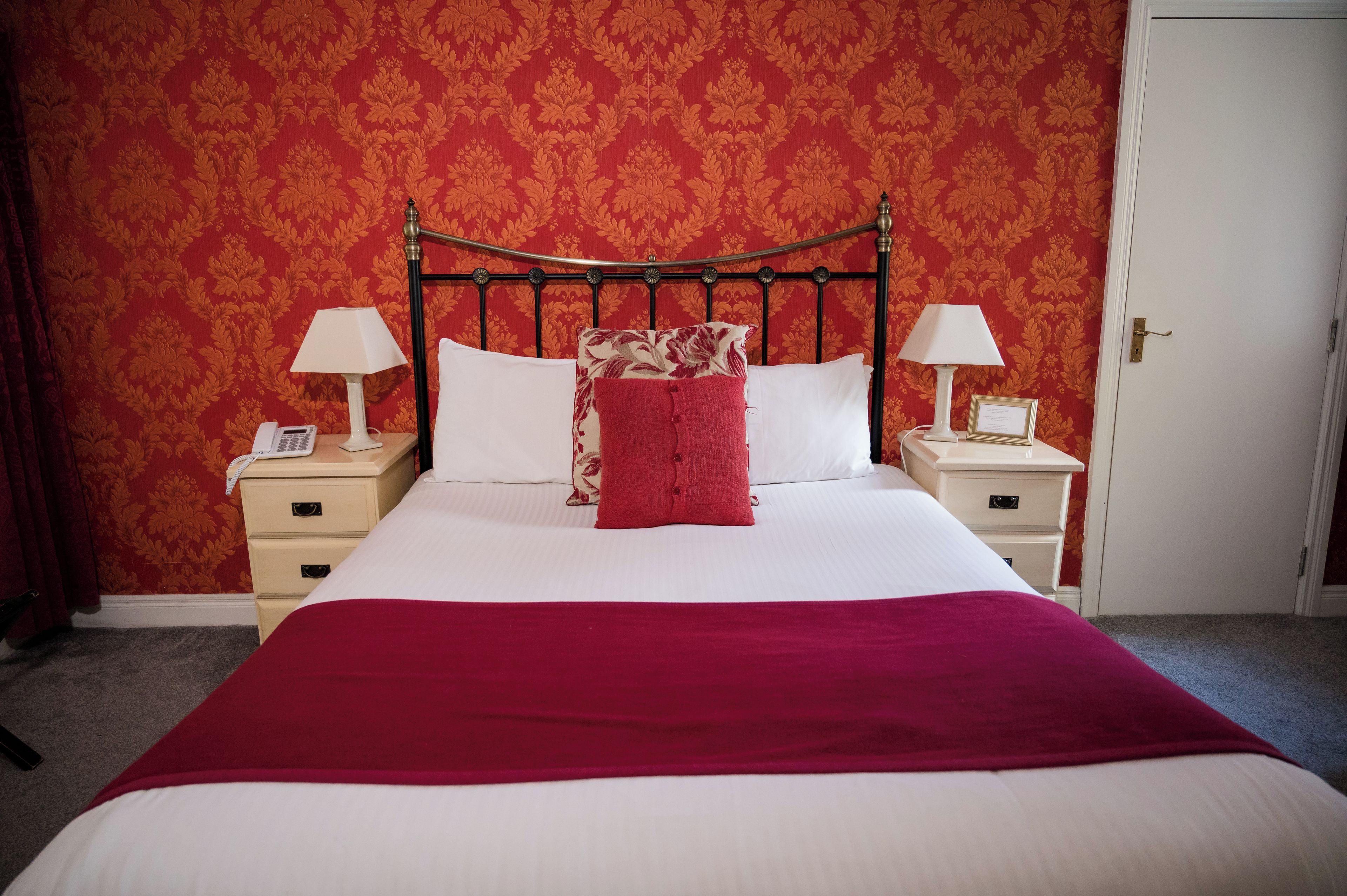 Una cama bien tendida con una colcha roja, dos lámparas de noche y un teléfono en la mesita de noche.