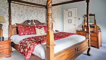 Una cama con dosel y cuatro columnas, con una colcha de estampado floral, una mesita de noche de madera con una lámpara, y un aparador con espejo.