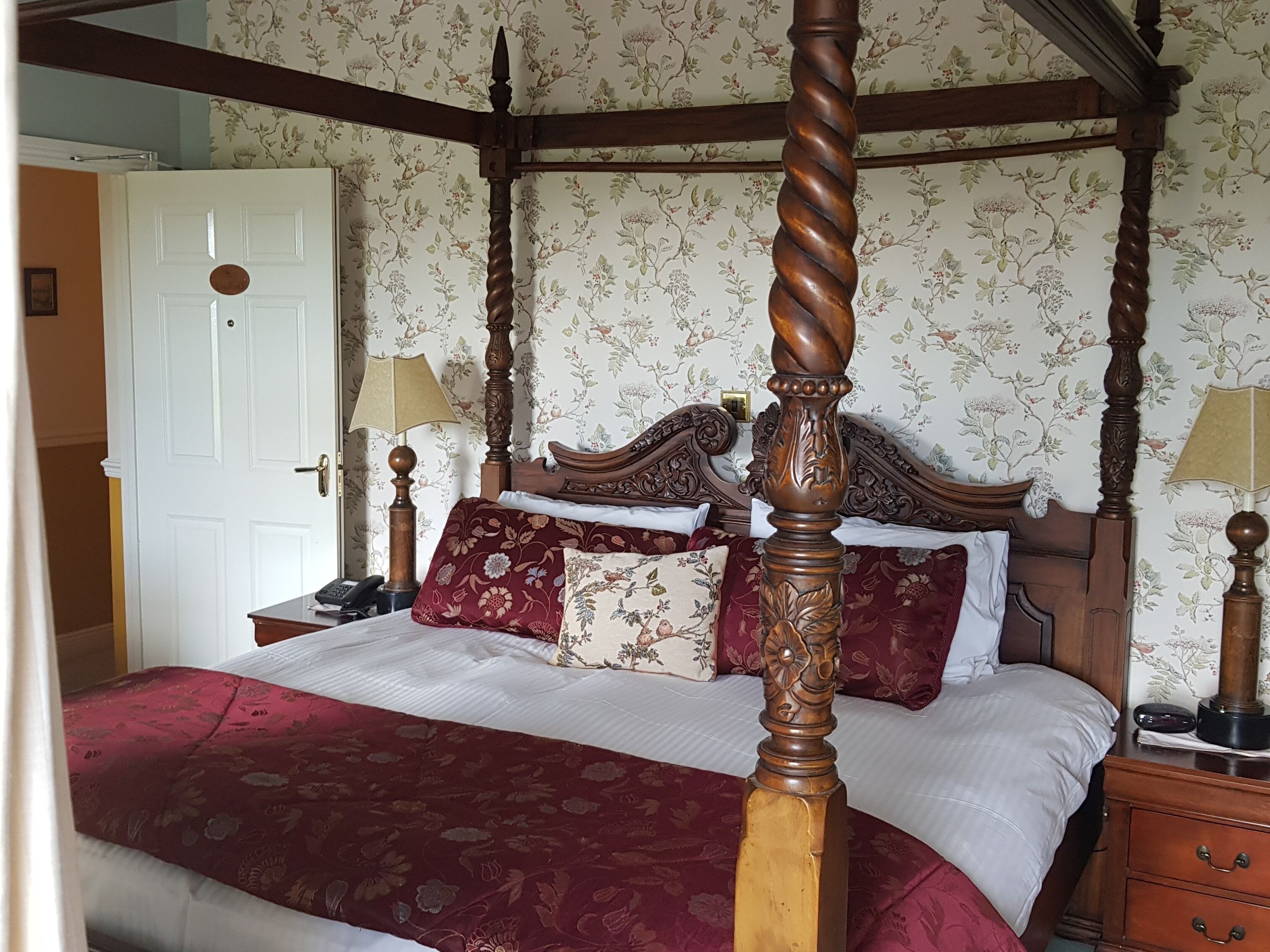 Uma cama com dossel, roupa de cama com padrão floral, cabeceira de madeira e mesa de cabeceira com abajur.