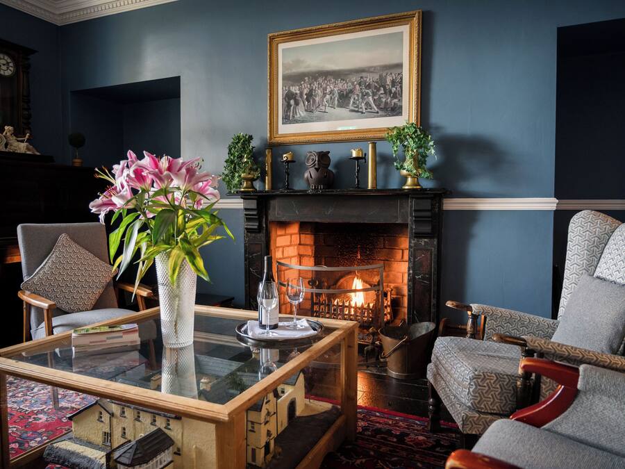 A cozy living room with a fireplace, comfortable seating, a glass coffee table, and a decorative vase with flowers.