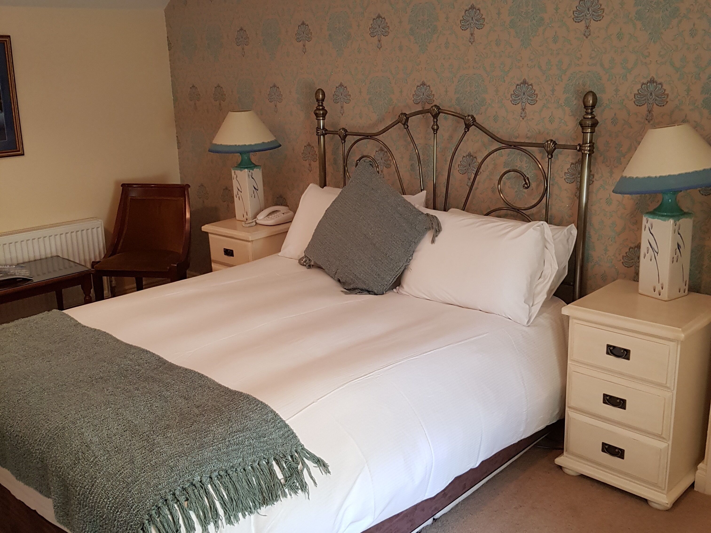 A neatly made bed with a grey throw, two bedside tables with lamps, a chair, and a radiator.