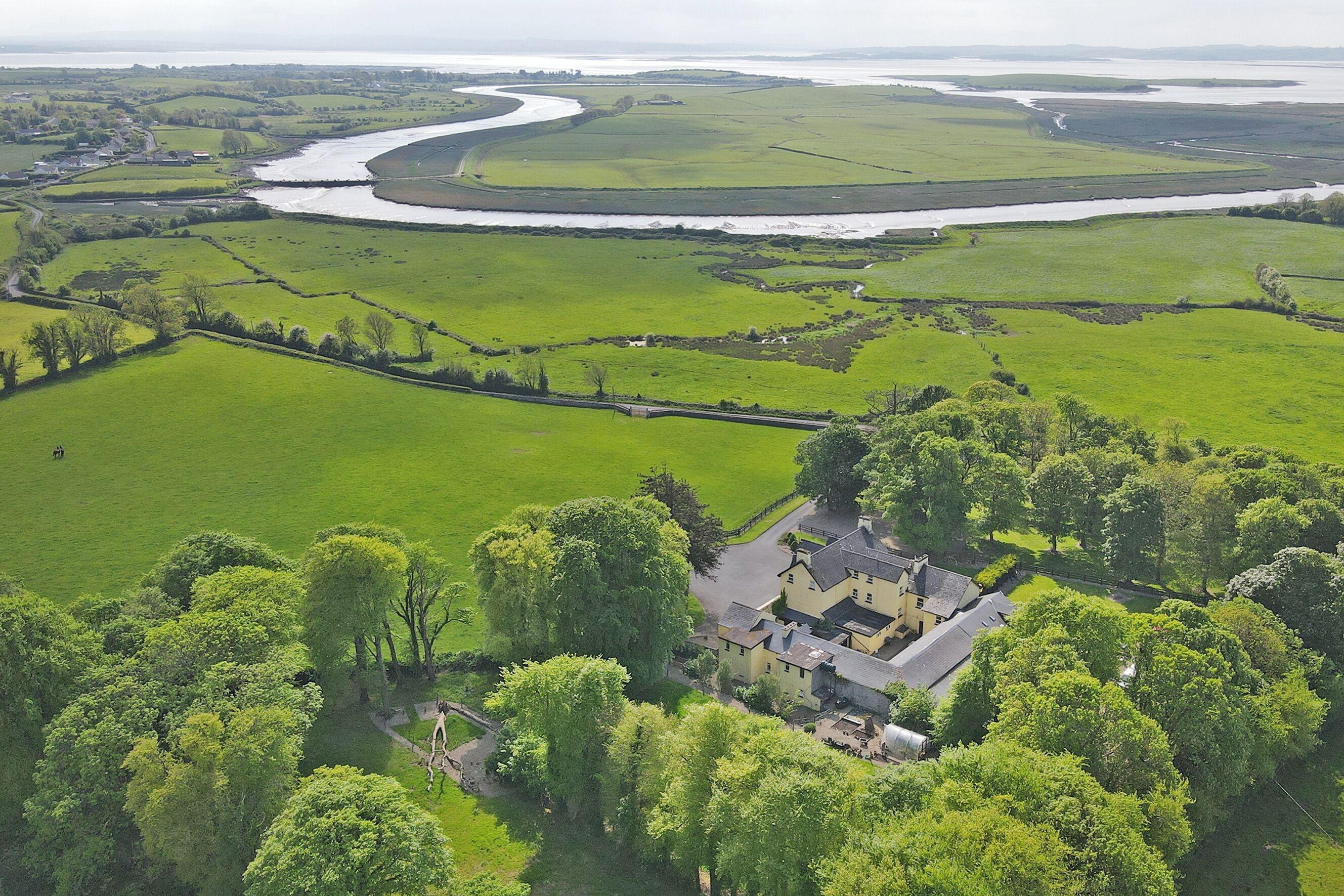 Une vue aérienne d’un domaine rural avec une grande maison entourée de champs verdoyants et traversée par une rivière sinueuse.