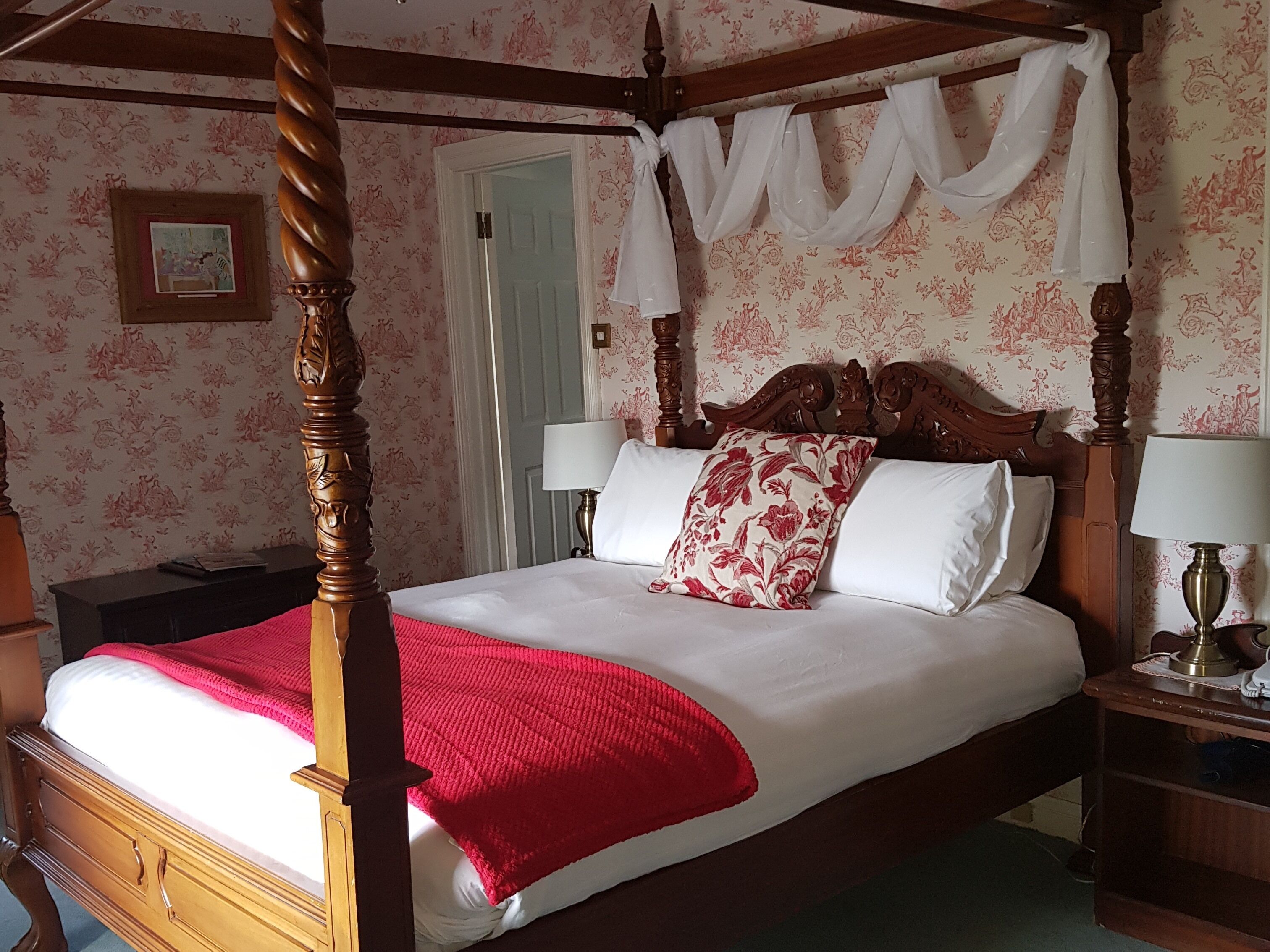 A four-poster bed with a red throw blanket, white linens, and floral pillows. There are two bedside tables with lamps. The room has floral wallpaper and a wooden ceiling.